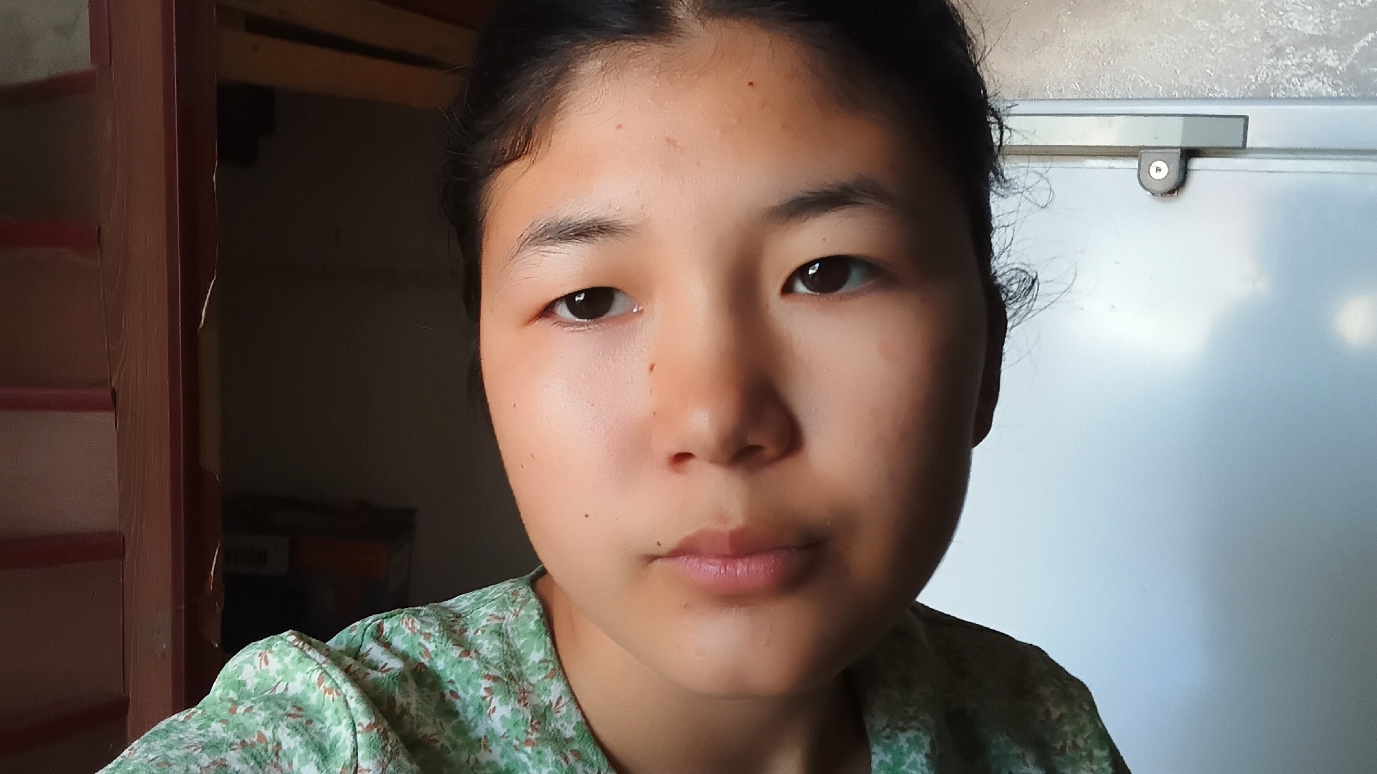 A young Asian woman is pictured in a close-up, frontal shot. She has dark hair pulled back and is wearing a green, floral-patterned shirt. Her skin is light with some freckles and a few blemishes on her forehead. Her eyes are dark and her lips are slightly parted. The background is softly blurred, with a wooden door frame on the left and a light-colored surface, possibly a refrigerator or whiteboard, on the right. The scene appears to be indoors.