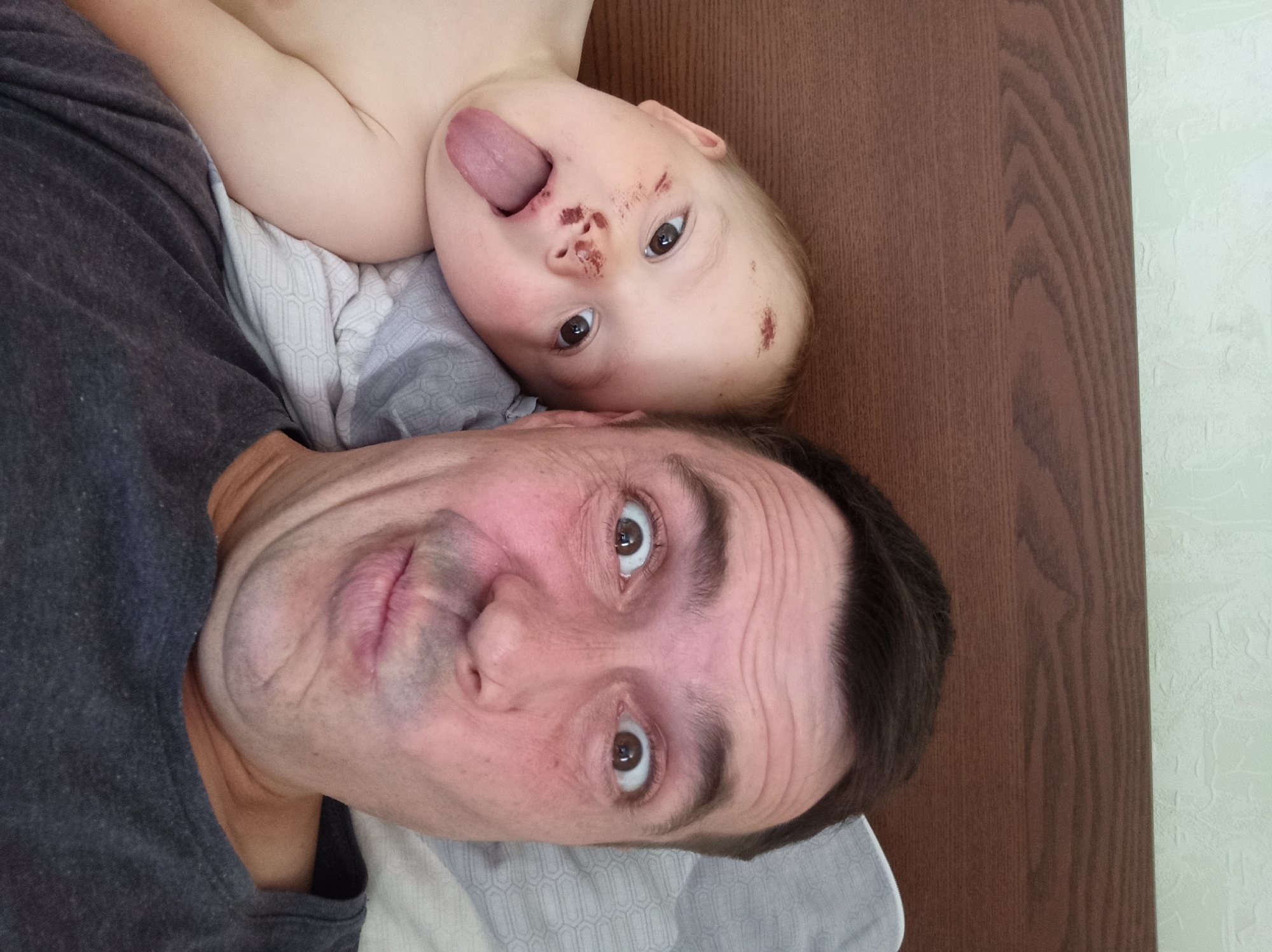 The image is a close-up, slightly tilted selfie taken by an adult male with a baby lying on his chest. The man is wearing a dark gray t-shirt. The baby is lying on its back, with its head resting on the man's chest and shoulder. The baby has its tongue sticking out and its eyes are open, looking towards the camera. The baby's face has some reddish marks, possibly from being wiped or from a minor injury. The man's face is also visible, with his eyes wide open and looking towards the camera. He appears to have wrinkles around his eyes. The background shows a wooden surface and a light-colored wall with a textured pattern. The setting appears to be indoors, possibly a bedroom, given the presence of what looks like a pillow or blanket beneath the baby. The lighting suggests it is daytime. There is no visible text in the image.