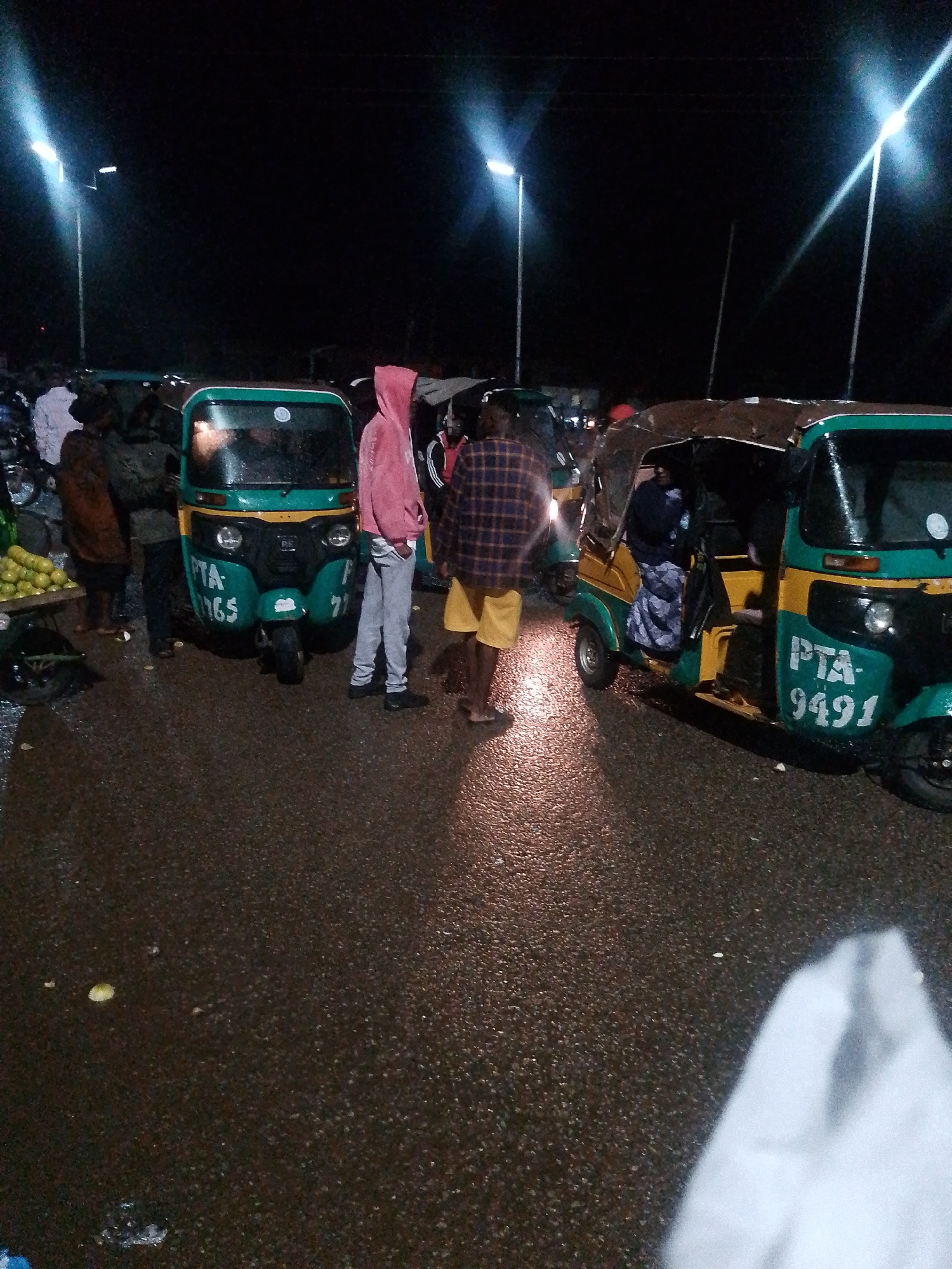 The image depicts an outdoor scene at night, likely in a city, with multiple auto-rickshaws visible. The ground is wet and reflective, suggesting it might be raining or recently rained. Several individuals are present, some standing near the auto-rickshaws and others appearing to be drivers or passengers. One auto-rickshaw in the foreground is green and yellow with "PTA 9491" written on its side. Another auto-rickshaw to the left is predominantly green with "PTA 1165" visible. People are dressed in casual clothing. The scene is illuminated by overhead streetlights, casting long shadows and creating a somewhat dimly lit atmosphere. The overall impression is of a transportation hub or a street corner where auto-rickshaws are operating at night.