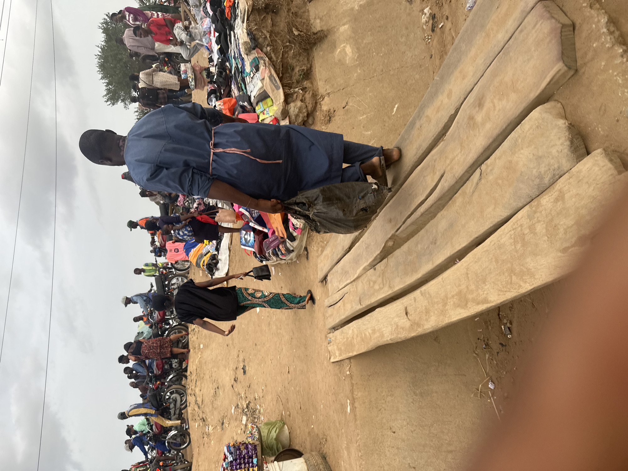 The scene depicts an outdoor market in Kuchingoro, Nigeria. In the foreground, there are four rough-hewn wooden planks. Adjacent to the planks, a woman in a black top and green patterned skirt walks away from the viewer, while a man in a blue uniform with a light pink tie carries a black plastic bag. Further into the scene, a group of people are congregating around merchandise, possibly clothing, displayed on a raised platform. In the background, a line of motorcycles is parked along a dirt road. The sky is overcast. A stand filled with assorted items sits next to a tire.