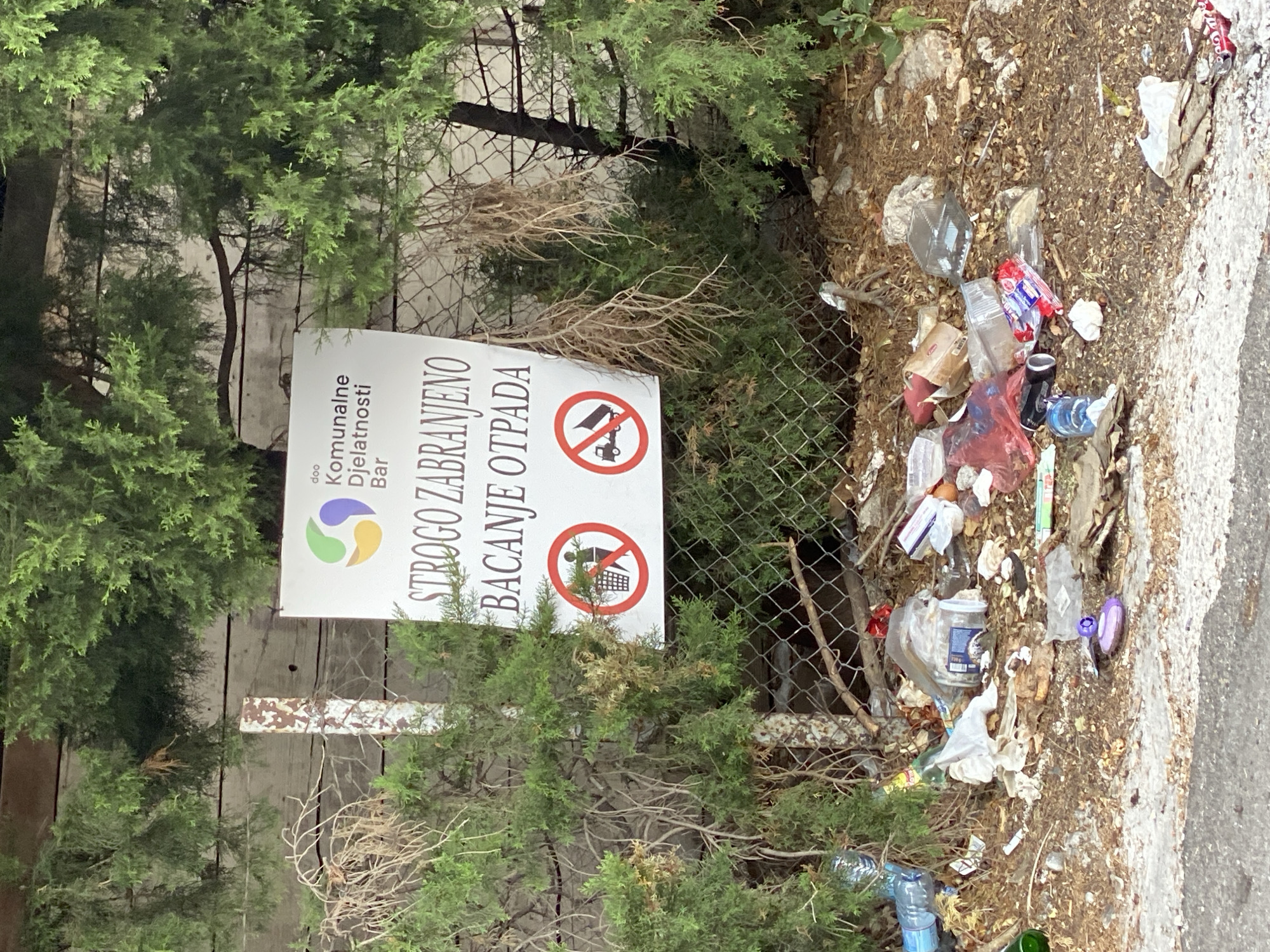 The image depicts a public area in Zgrade, Montenegro, where a sign is posted with the text "STROGO ZABRANJENO BACANJE OTPADA," meaning "Strictly Forbidden to Throw Trash." The sign also features two prohibition symbols: one showing a person throwing trash into a bin with a cross over it, and another showing a truck dumping garbage with a cross over it. The area around the sign is heavily littered with various types of waste, including plastic bottles, bags, food wrappers, and other debris. Green foliage, likely trees or bushes, frames the scene, and a chain-link fence separates the littered area from what appears to be a path or road in the foreground. The overall impression is one of neglect and improper waste disposal despite the explicit warning sign.