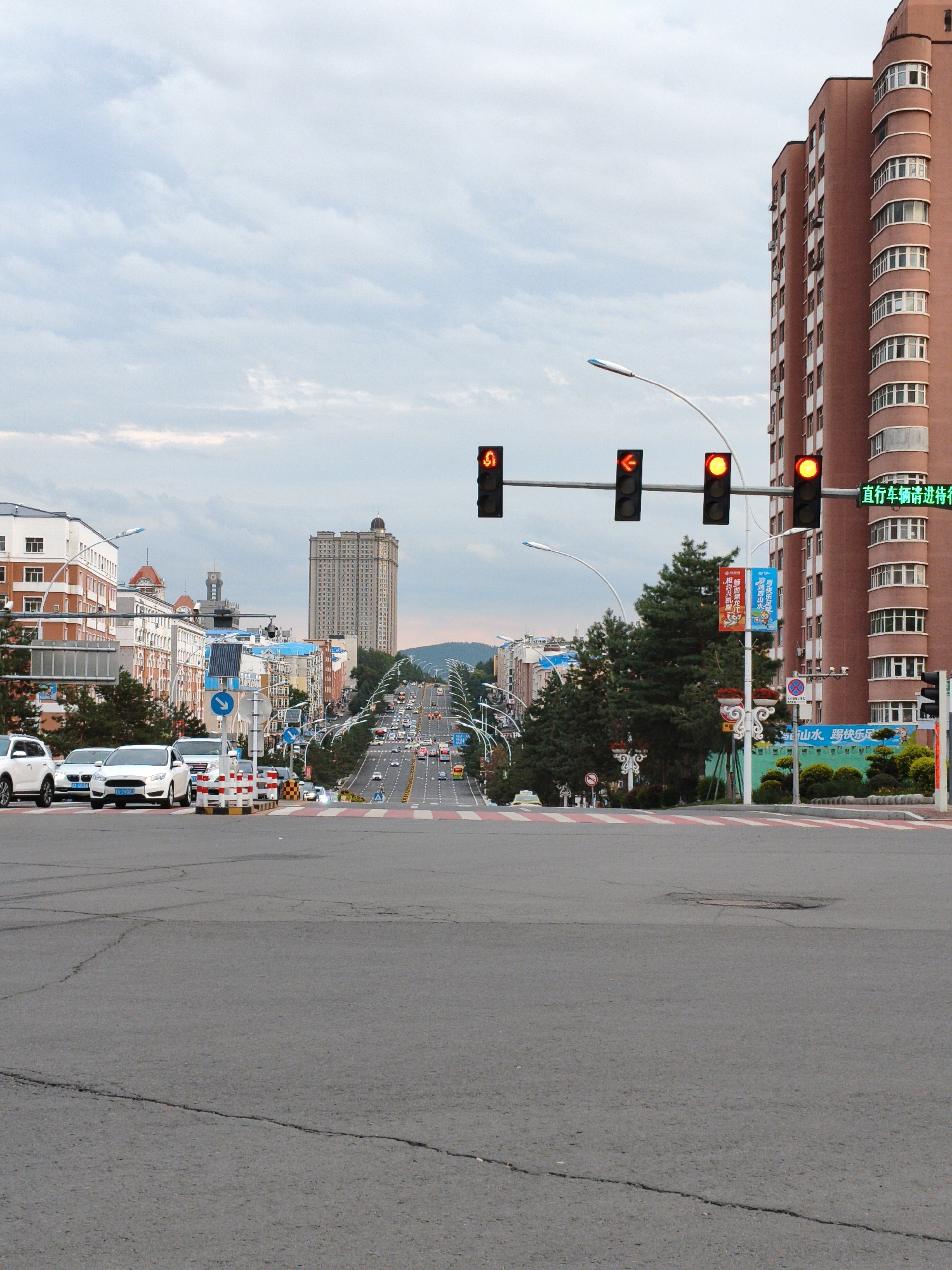 The image shows a street view in Jixi, China, likely during the daytime with overcast skies. The foreground features a wide asphalt road with a marked pedestrian crossing in red and white stripes. A few cars are stopped at the crossing. In the background, a long, uphill road stretches into the distance, lined with buildings on either side and trees. Traffic lights are visible, with some showing red. The architecture includes modern apartment buildings and older structures. The overall scene depicts a typical urban street in a Chinese city, with elements of urban infrastructure and daily life.