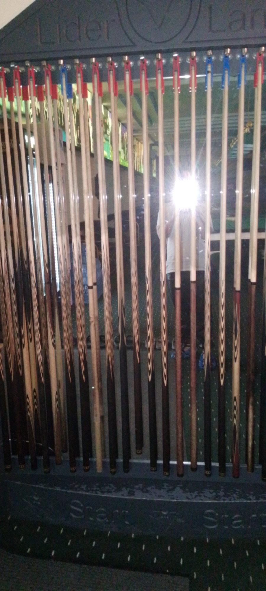 The image displays a collection of billiard cues arranged vertically in a rack. The rack is dark gray and has "Lider" and "Lan" inscribed at the top, suggesting a brand or model name. Below the inscription, there are colorful clips, some red and some blue, holding the cues in place. The cues themselves vary in design, with some appearing to be made of light-colored wood, while others have dark wood or intricate striped patterns. In the background, a mirror reflects a person standing in front of it, their face obscured by a bright light. The overall environment appears to be an indoor space, possibly a billiard hall or a storage area for sports equipment. The image does not provide specific visual cues to determine the location within the city of Angren, Uzbekistan.