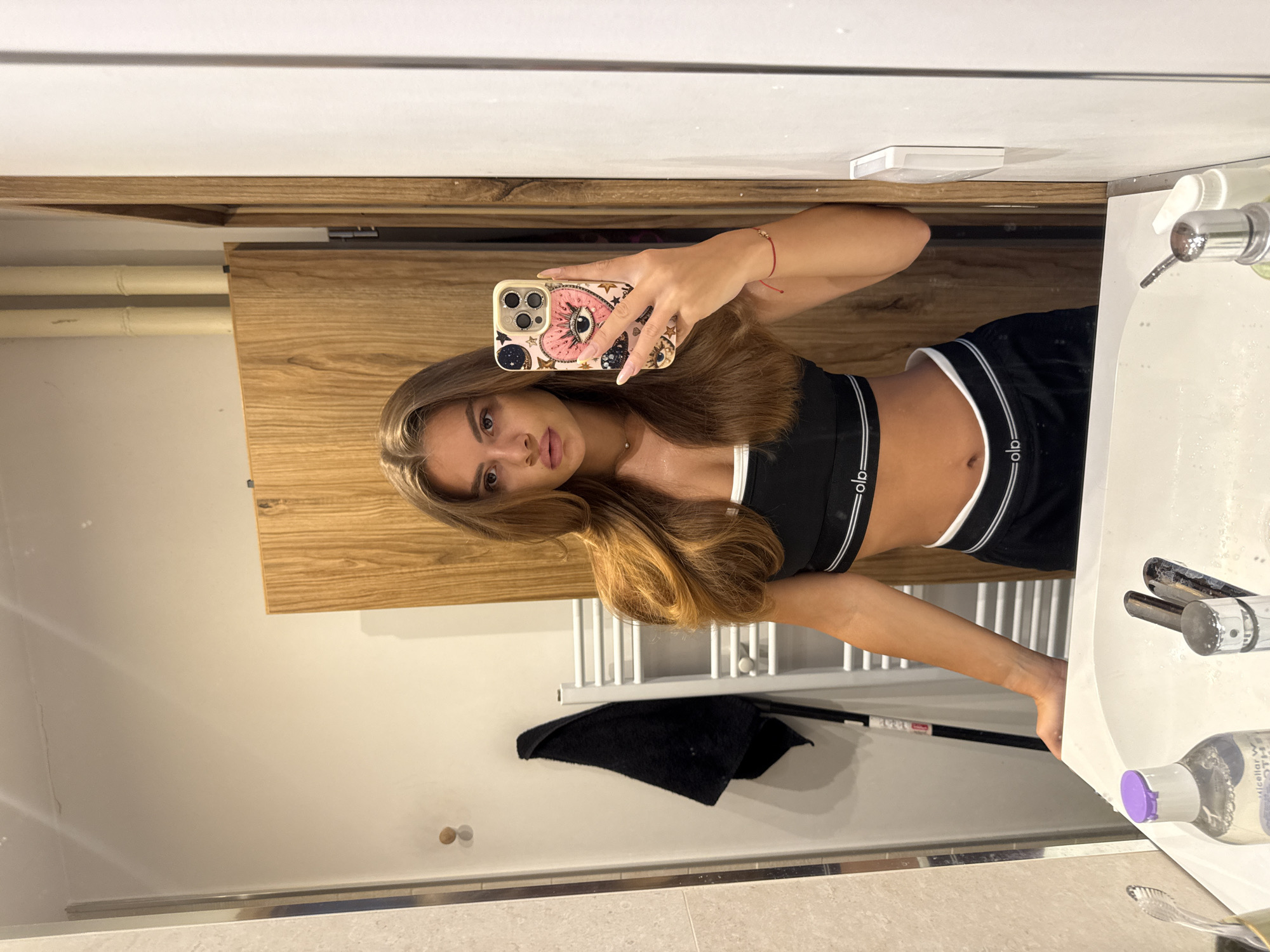 A young woman with long, auburn hair takes a mirror selfie in a modern bathroom. She is wearing a black sports bra and matching shorts, both with white trim and branding. Her left hand is holding an iPhone with a decorative case featuring eyes and celestial motifs, while her right arm rests on the sink. The bathroom features light-colored tiled walls, a wooden cabinet, and a white sink with chrome fixtures. A black towel hangs on a heated rack in the background. The image was taken in Plovdiv, Bulgaria, within a residential or hotel bathroom.