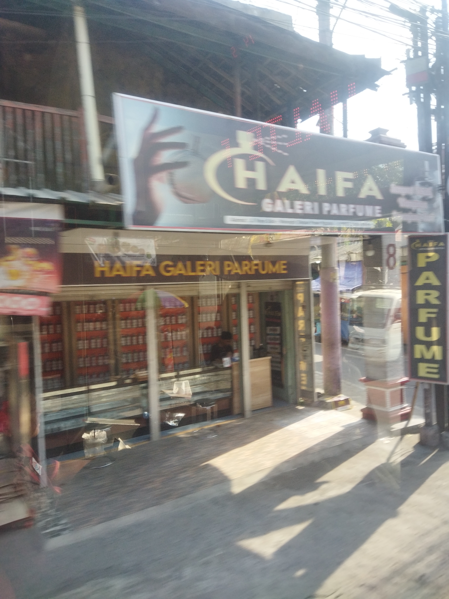 In the city of Nguter, Indonesia, a storefront for "HAIFA GALERI PARFUME" is captured from a slightly elevated, moving perspective. The facade of the shop features large glass windows displaying rows of perfume bottles, predominantly in red and white packaging, organized on shelves. Above the storefront, a prominent sign with the text "HAIFA GALERI PARFUME" in yellow lettering is displayed on a dark background. Another vertical sign on the right side of the image also reads "PARFUME" in yellow letters. The street in front of the shop is paved and shows long shadows cast by an overhead structure, likely a roof or awning. The scene is illuminated by bright sunlight, suggesting it is daytime. There are glimpses of people inside the store, including one individual behind a counter. The overall impression is of a commercial establishment selling perfumes.