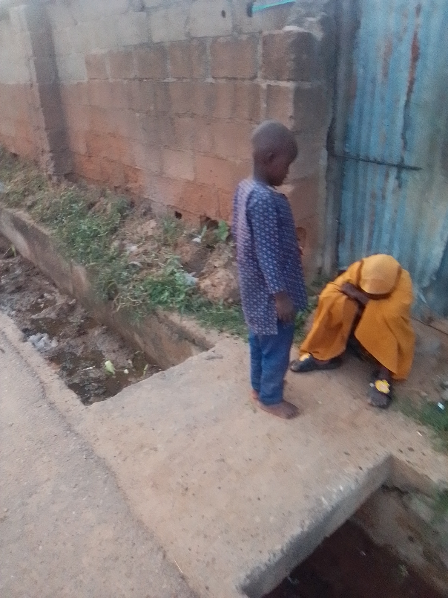 Two young children are outdoors in an urban environment. One child, wearing a blue patterned shirt and blue pants, stands facing away from the camera and looking down. The other child, dressed in a yellow garment that covers their head and body, is crouched on the ground with their arms wrapped around their legs, appearing distressed. They are situated next to a concrete drainage ditch filled with murky water and debris. Behind them, a brick wall and a corrugated metal door are visible. The ground is a mix of concrete and dirt. The scene appears to be located in Liji Hill, Nigeria, in a somewhat dilapidated area with evidence of poor sanitation.