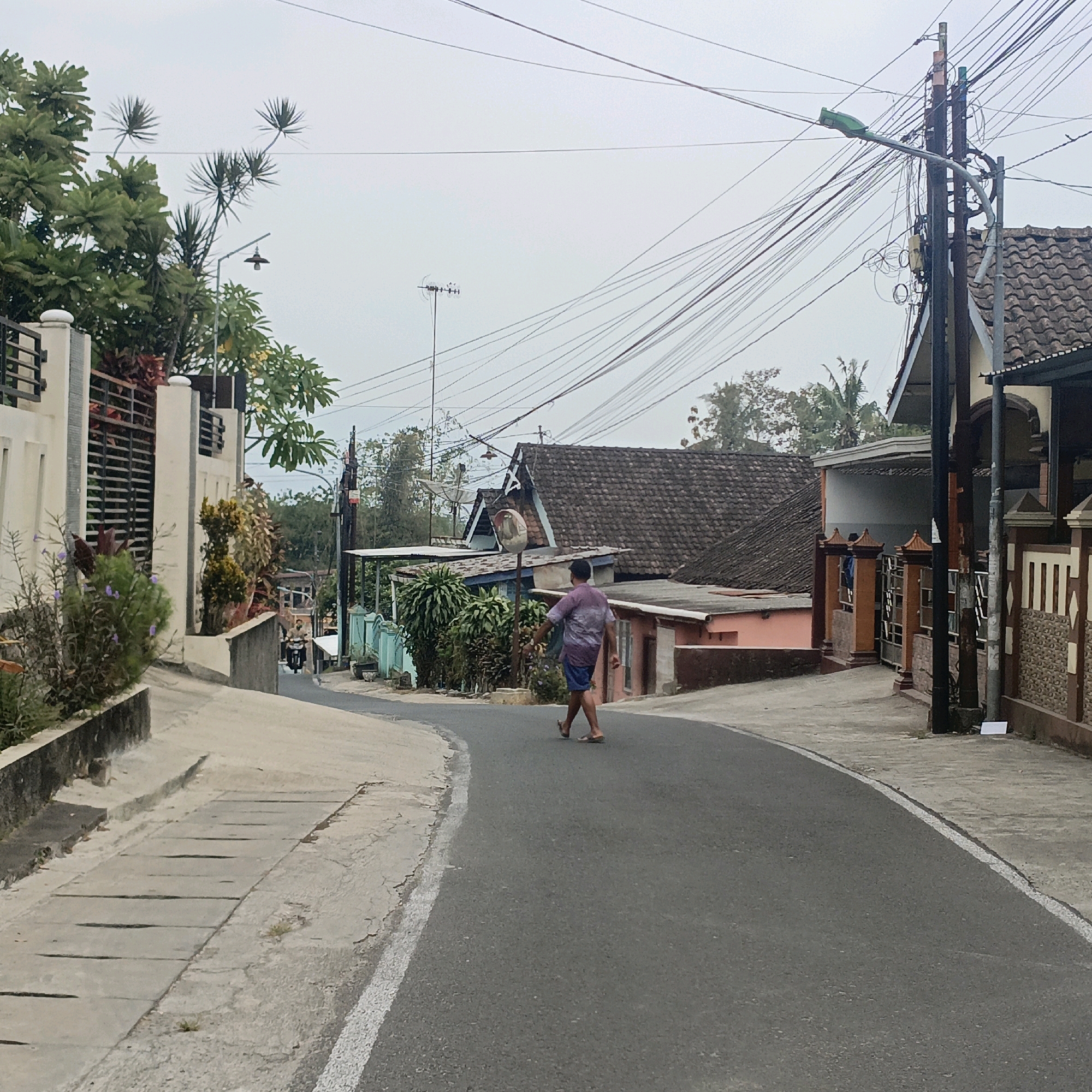 The image depicts a street scene in Salatiga, Indonesia, on an overcast day. A man in a purple tie-dye shirt and blue shorts is walking away from the camera on the right side of the road. The road curves to the left and leads uphill. On the left side of the road, a concrete sidewalk runs alongside a wall with a black metal fence and lush green plants. Houses with tiled roofs and visible electrical wires are situated on both sides of the street, with some structures featuring wooden and tiled facades. A utility pole with numerous tangled wires stands prominently on the right side of the road. In the distance, a person on a motorcycle is visible. The overall atmosphere suggests a residential area in a tropical climate.