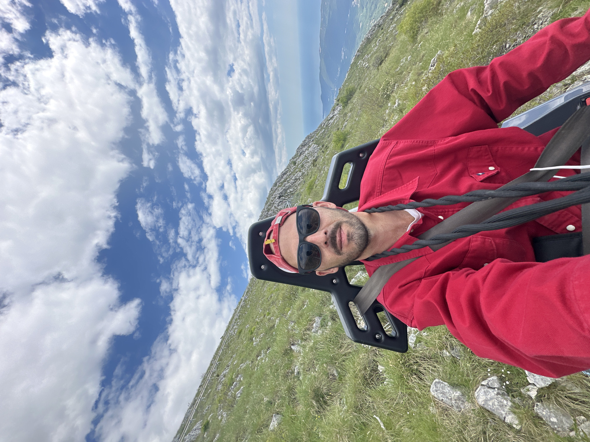 A man wearing sunglasses and a red jacket sits in a ride with a black harness, possibly a zipline, in an outdoor setting. Patches of grass and rocky terrain are visible below, while a body of water and cloudy sky are in the background. Based on the context, the location is likely near Radanovići, Montenegro, given the user's query.