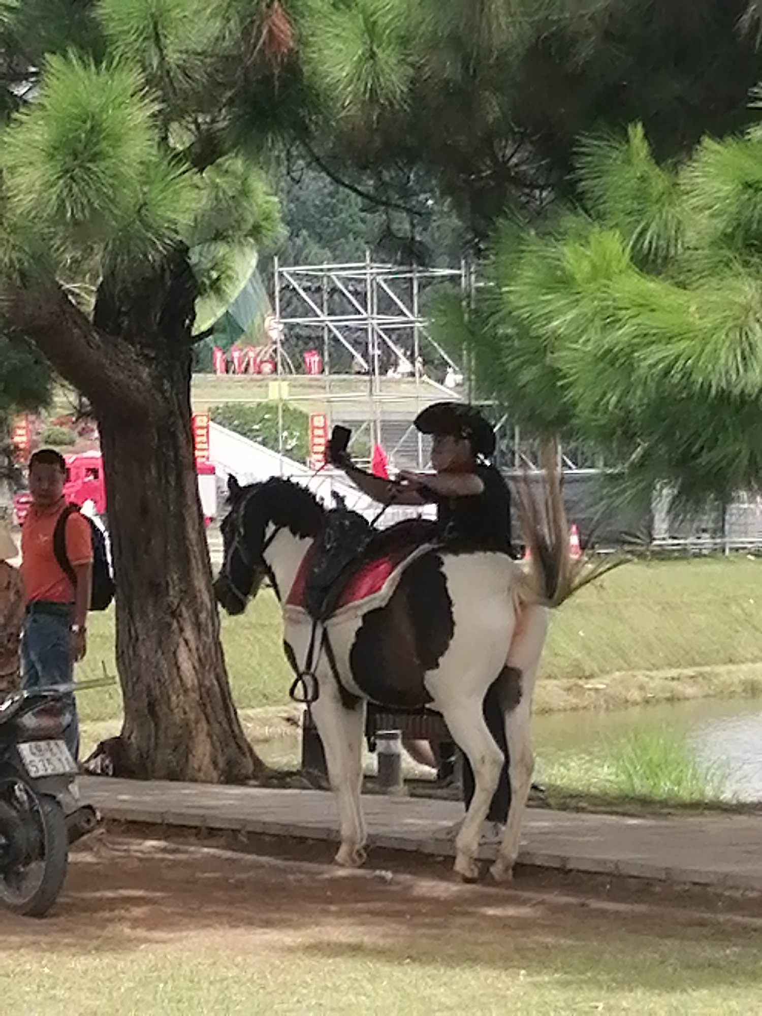 The image depicts a scene in Da Lat, Vietnam, where a person is riding a piebald horse. The rider, wearing a dark top and a cowboy hat, is holding up a smartphone, seemingly taking a selfie or recording a video. The horse is equipped with a saddle and bridle. To the left, a man wearing an orange polo shirt and jeans stands near a tree, with a backpack slung over his shoulder. A motorcycle is parked to the far left of the frame. In the background, scaffolding structures and red flags suggest an event or construction is taking place. The environment appears to be outdoors, with lush green trees and a body of water visible. The overall lighting indicates it is daytime.
