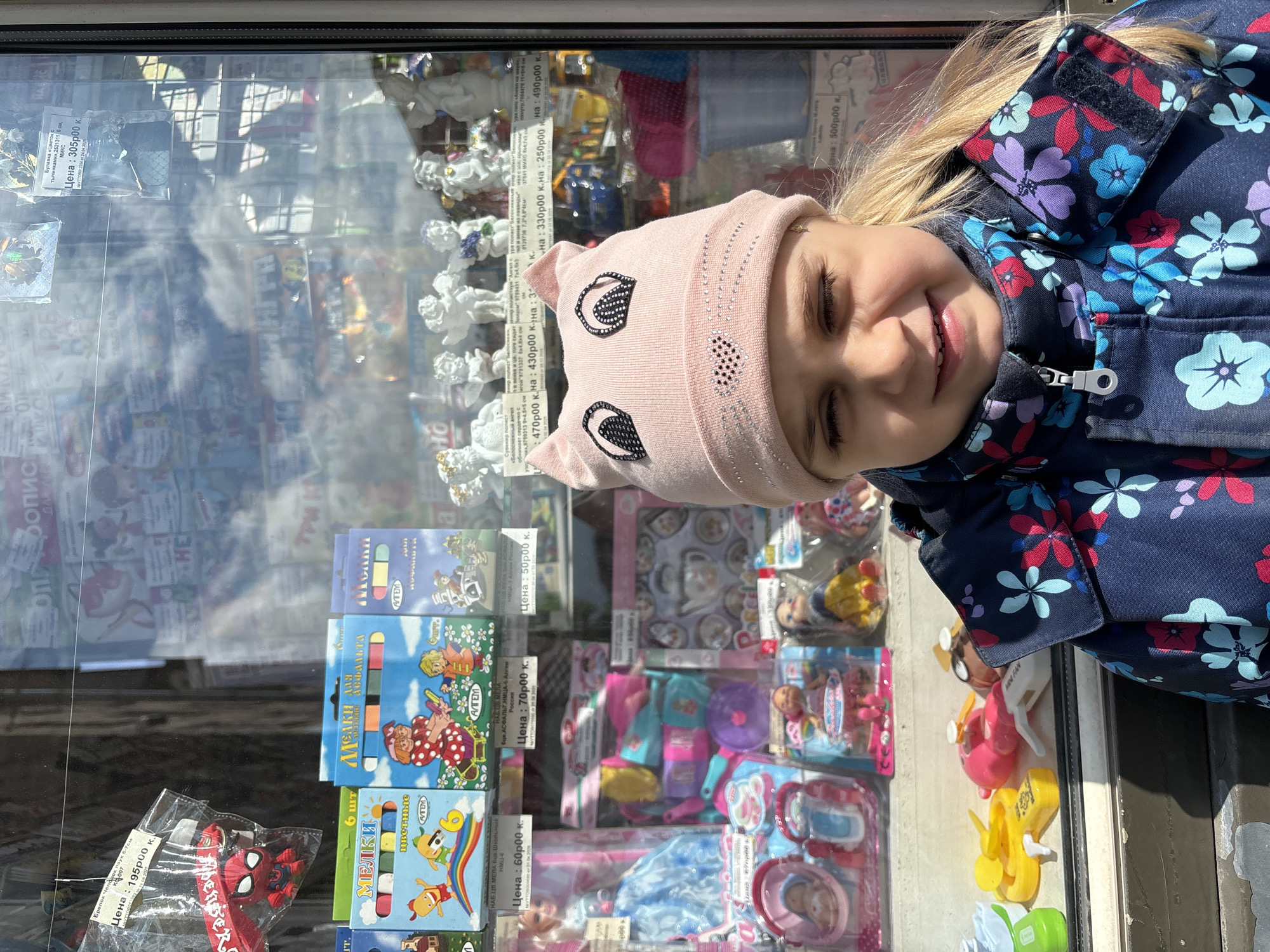 A young girl stands in front of a shop window filled with toys and other items for sale in Saint Petersburg, Russia. The girl is wearing a pink cat-eared hat and a floral-patterned jacket. She is smiling. The shop window displays a variety of items, including chalk, toys, and decorative figurines. Prices are marked in rubles. The reflection in the window suggests the presence of buildings across the street, though the image is distorted. The lighting indicates it is a sunny day.