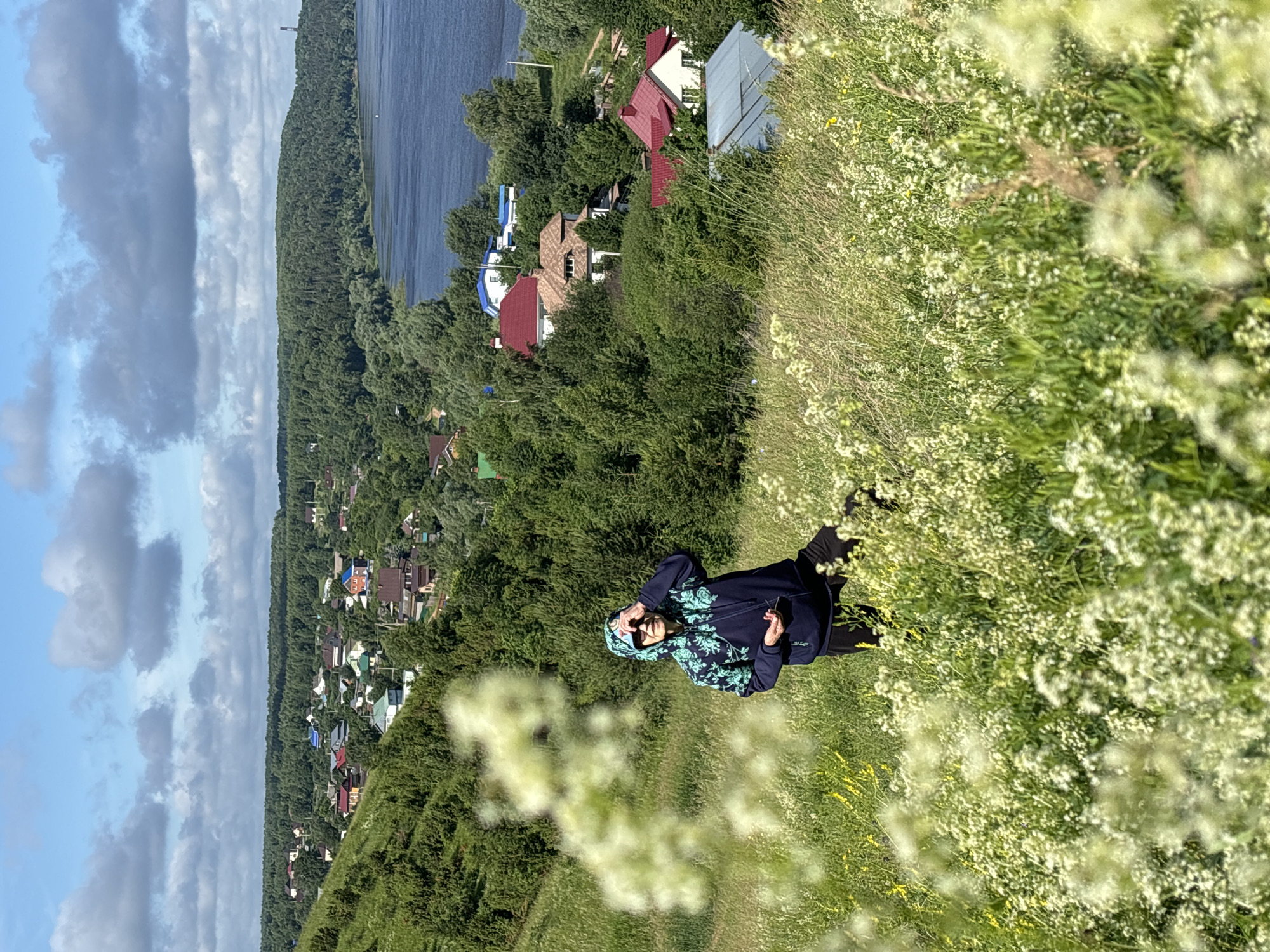 In the heart of Olkhovka, Russia, a young woman is captured in a moment of serene enjoyment amidst a vibrant natural landscape. She's dressed in a dark blue hooded jacket adorned with a light green floral pattern and black leggings, her gaze directed downwards as she holds a phone in her right hand. Her presence is set against a backdrop of rolling green hills, abundant with white wildflowers and scattered yellow blooms, suggesting a late spring or early summer day.
The scene unfolds overlooking a picturesque village nestled amongst lush green trees, with the tranquil blue waters of a river or lake in the distance. Several houses with colorful roofs, varying in shades of red, blue, and grey, are visible, hinting at a peaceful rural community. The sky above is a canvas of soft blue, punctuated by scattered white clouds, indicating fair weather.
The overall mood conveyed is one of tranquility and appreciation for nature's beauty. The soft sunlight suggests it is likely midday or early afternoon, further enhancing the peaceful atmosphere. The image is oriented vertically, with the woman positioned on the right side of the frame, looking out over the expansive landscape.