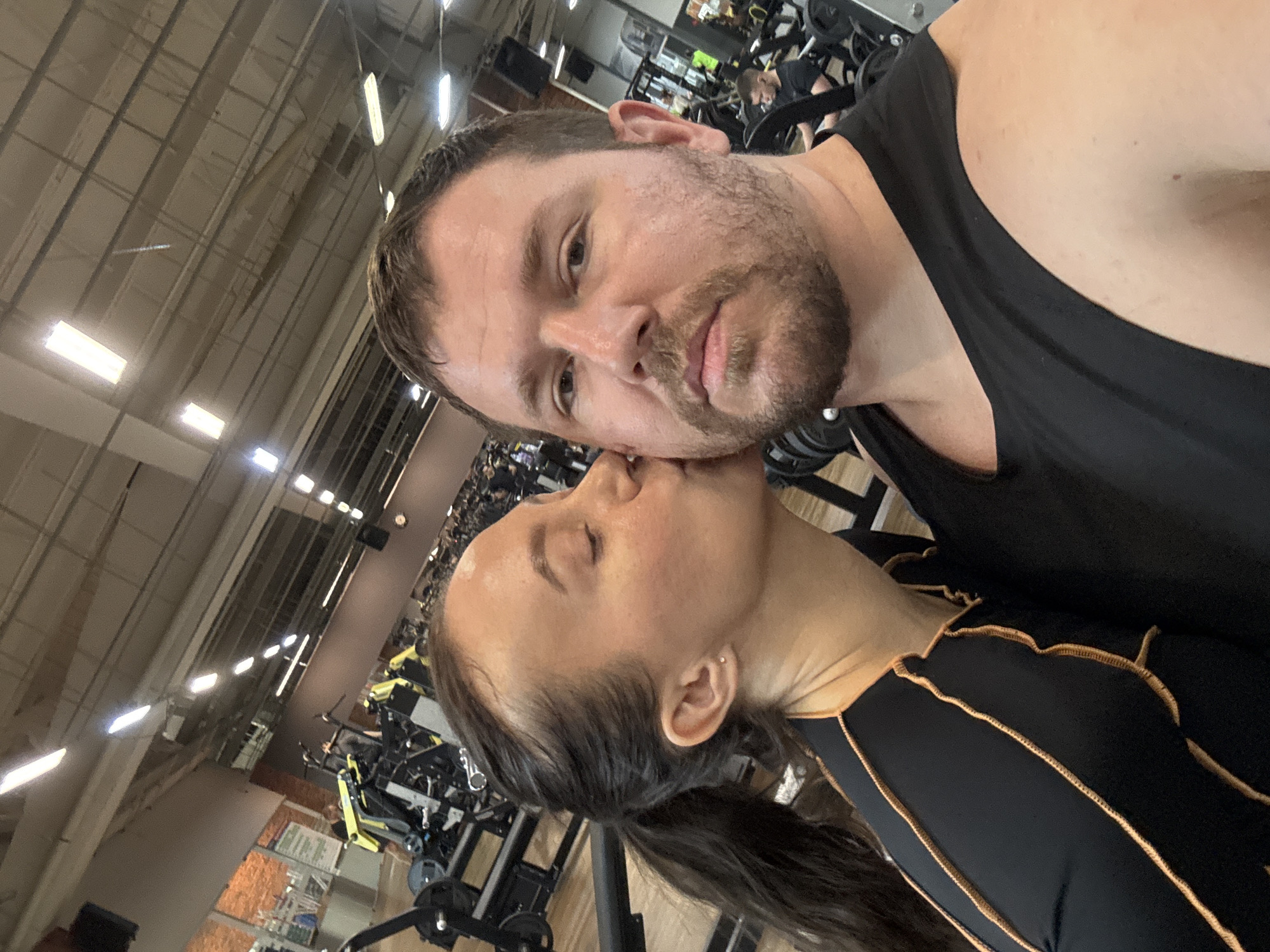 The image depicts a man and a woman in a gym setting. The man, in a black tank top, is looking directly at the camera. The woman, wearing a black athletic top with gold trim, is kissing his cheek. Gym equipment and other individuals are visible in the background. The image was taken in Chaikovsky, Russia.