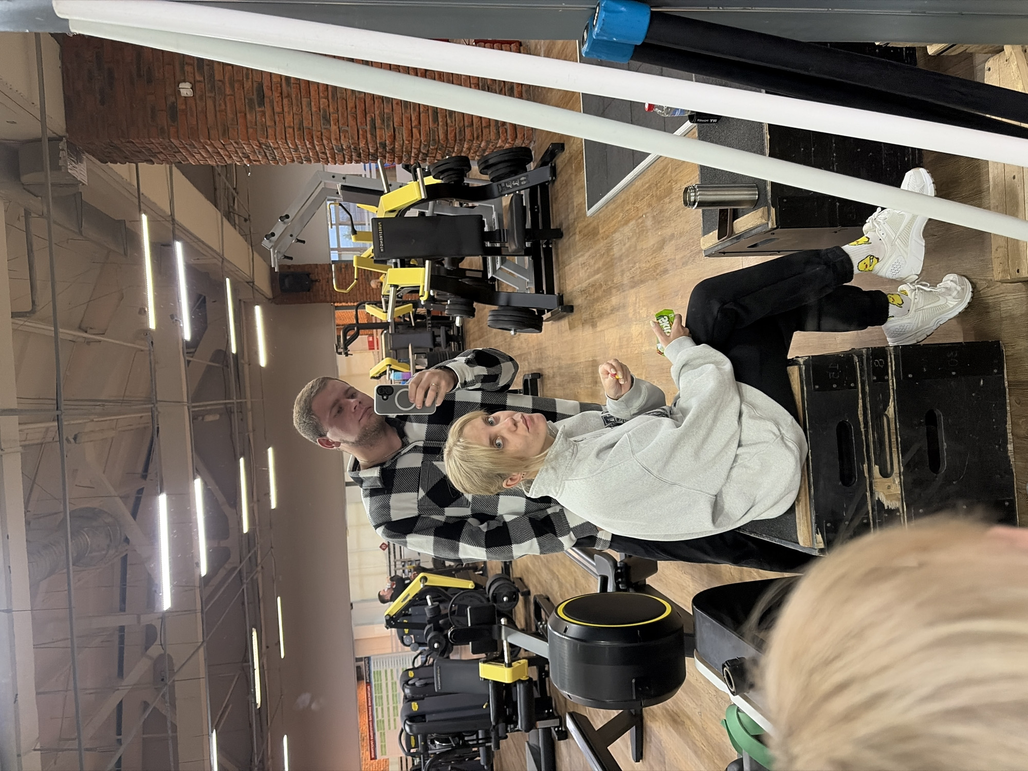 A man and a woman are posing for a selfie in a gym, likely in Chaikovsky, Russia, given the building architecture and interior design style. The man is holding a smartphone and wearing a black and white checkered shirt. The woman is sitting on a piece of gym equipment, wearing a grey hoodie and black pants. She is holding a small container in her hand. Various gym equipment are visible in the background, including rowing machines and weightlifting apparatus. The gym has a high ceiling with fluorescent lighting, and a brick wall is visible in the upper left corner.