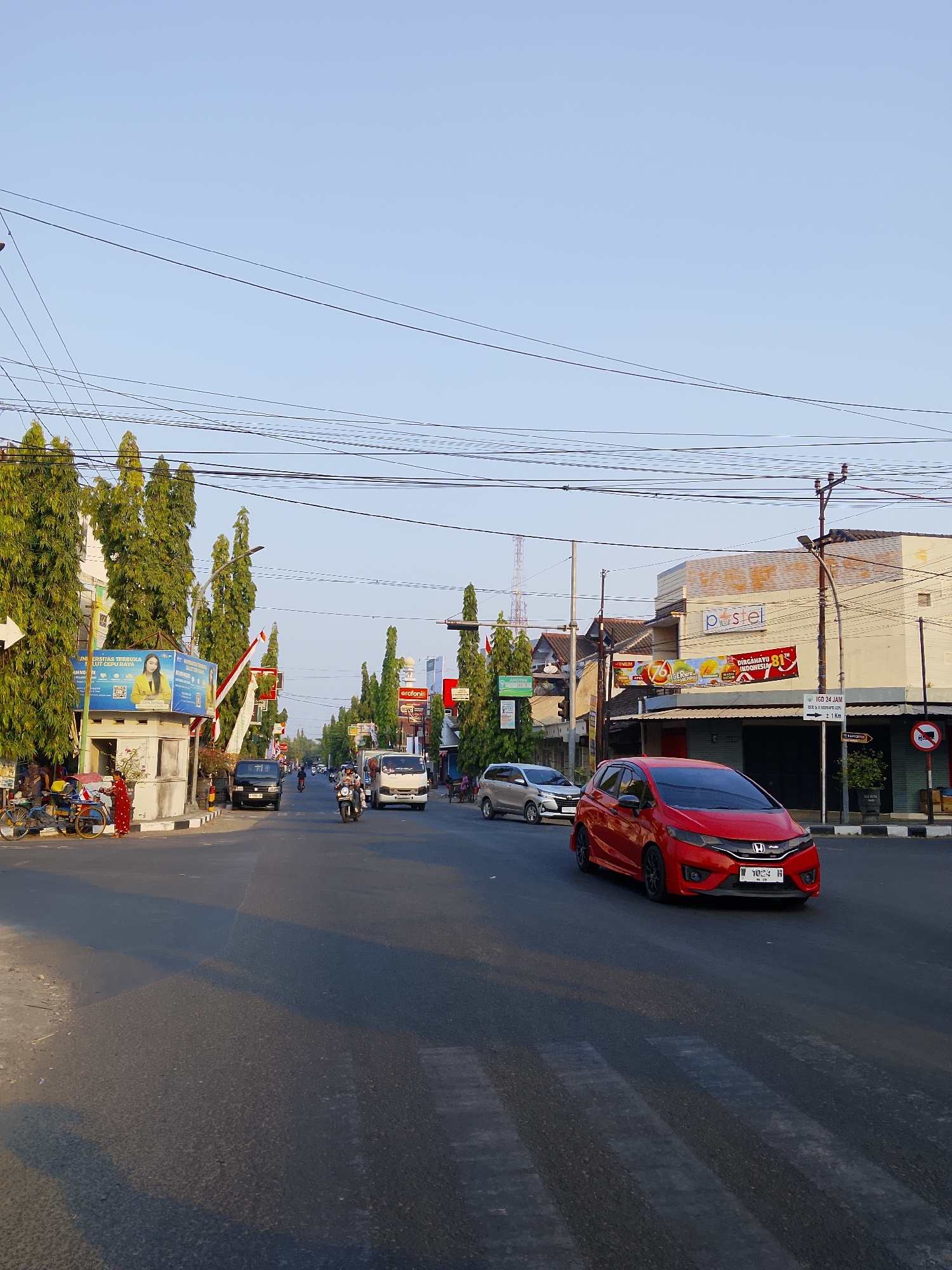 The image depicts a street scene in Sambong, Indonesia, likely during the daytime given the clear sky and shadows. The street is paved and appears to be a main thoroughfare with multiple lanes of traffic. Overhead power lines crisscross the sky, supported by poles.
On the left side of the road, there are several tall, slender trees with dense green foliage, lining the sidewalk. Adjacent to the trees is a building with a white facade and a large blue billboard featuring a woman and text in Indonesian. A small, grey booth-like structure is situated in front of the building. A bicycle and a pedicab are parked near the sidewalk.
The road itself is populated with various vehicles. A red hatchback car is prominently featured in the foreground, traveling on the right lane. Its license plate is partially visible. To the left of the red car, a silver hatchback is also in motion. Further down the road, a black van and a white delivery truck are visible. Several motorcycles are also present, with one in the middle of the road appearing to be ridden by two people. A pedestrian in a red dress is standing near the sidewalk on the left.
On the right side of the street, there are buildings with various signs. One building has a sign that reads "pastèl" in pastel colors. Another sign displays a promotional advertisement with an image of fruit. Further down, a sign indicates "IGD 24 JAM" and an arrow pointing to the right. A traffic sign with a red circle and a diagonal bar indicating a prohibition is also visible. The environment appears urban, with buildings, road infrastructure, and vegetation. The overall impression is that of a busy street in a residential or commercial area.