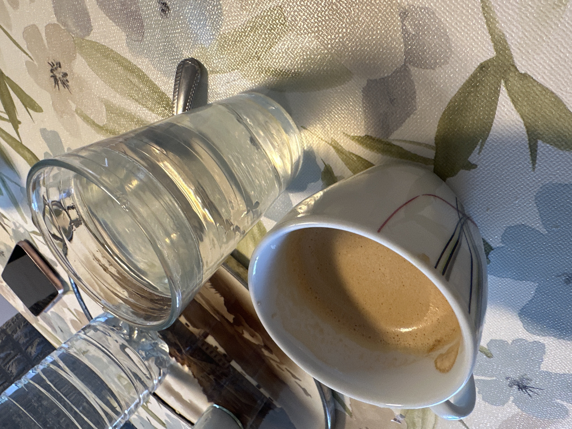 This image, taken in Mainburg, Germany, captures a still life of beverages and personal items on a patterned surface. A clear glass tumbler, filled with water, occupies the central left of the frame. To its right, a white ceramic coffee cup, containing a light brown beverage with foam (likely coffee or espresso), is visible. The cup features thin, multi-colored vertical lines in red, blue, yellow, and black on its exterior. A silver spoon is partially visible behind the glass.
In the lower left corner, a black smartphone with a metallic bezel is partially visible, next to a clear plastic water bottle with horizontal ridges. All these objects rest on a light-colored tablecloth or placemat with a floral pattern. The pattern includes light grey, purple, and blue flower blossoms, along with green leaf designs, against a textured background. The lighting is soft and even, indicating an indoor setting, possibly during the day. No people are present, and there is no visible text on any objects. The scene depicts a quiet moment, perhaps before or after a meal or break.