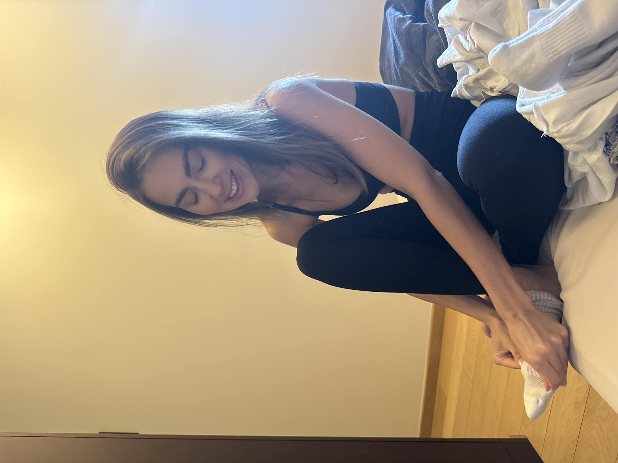 A woman is sitting on a bed. She is wearing a black sports bra and leggings, and she is putting on a white sock. There are clothes piled next to her on the bed. The scene is in a room with light yellow walls and light wood trim. A piece of furniture is visible in the foreground. The location is in the city of Unknown, Unknown.