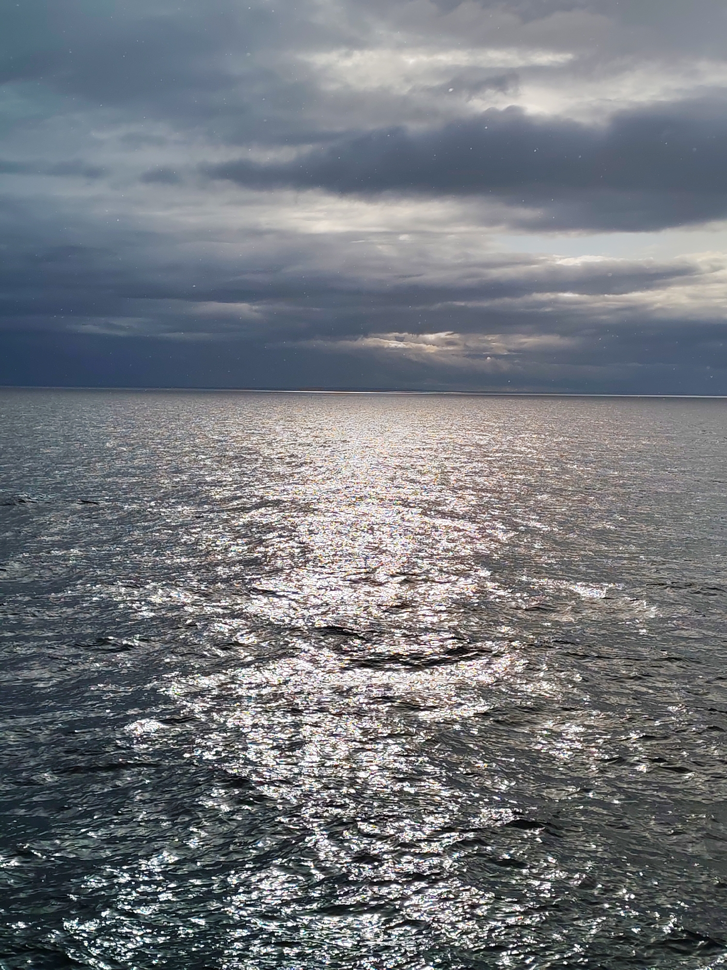The image depicts a body of water under an overcast sky. The water's surface reflects sunlight, creating a shimmering effect. Dark, heavy clouds dominate the sky, with a small break allowing some light to penetrate. The location is Unknown, Russia. No people or specific objects are visible.