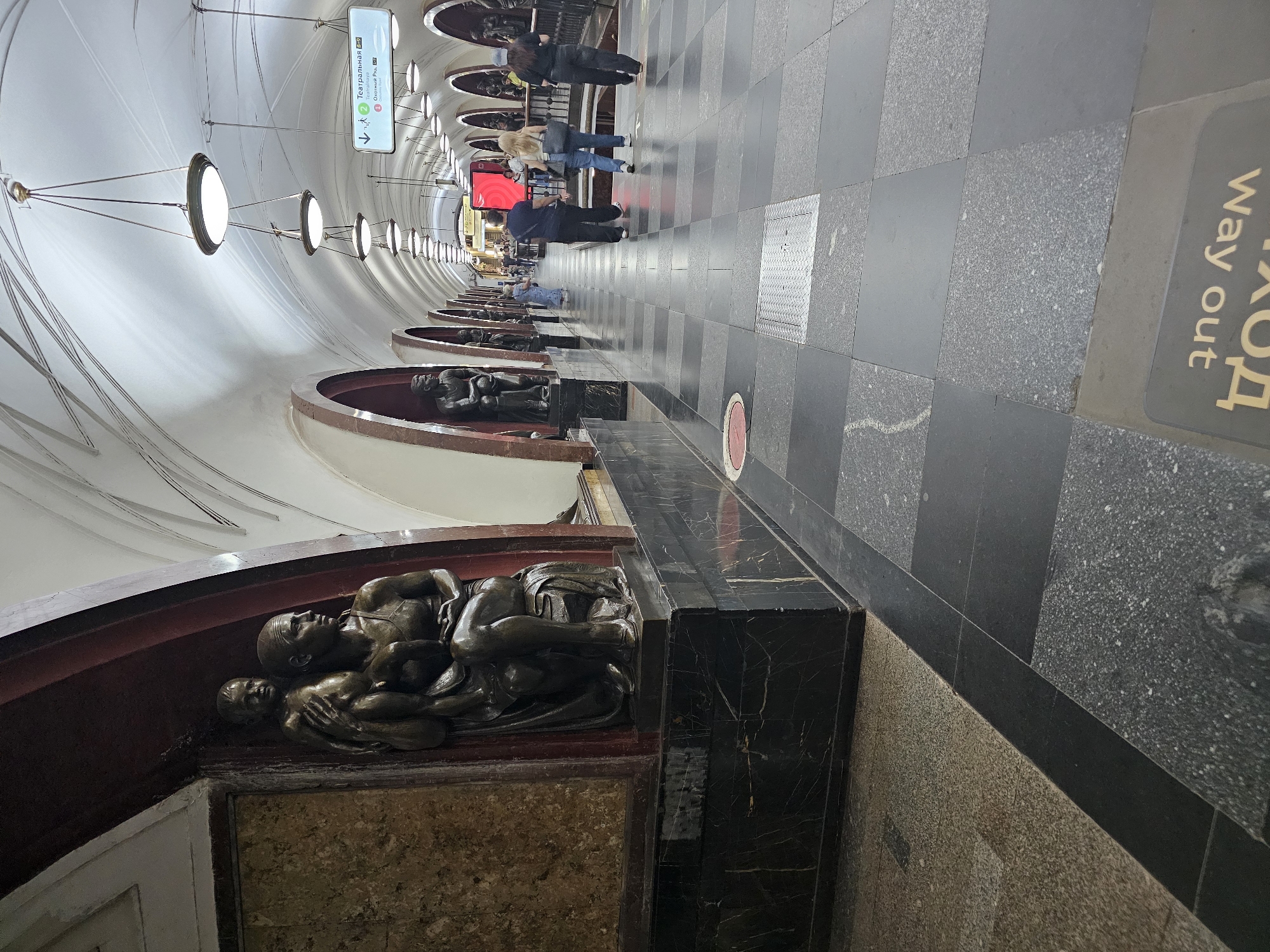 This image depicts the interior of a Moscow Metro station, likely Sokolnicheskaya Line based on the architectural style and signage. In the foreground, a large bronze sculpture of a woman holding a child is prominently featured on the left. The sculpture is set within an arched alcove, accented by dark marble and patterned stone. The station features a long, vaulted ceiling with evenly spaced pendant lights. Along the platform, several other sculptures are visible within similar arched recesses, creating a symmetrical visual rhythm. People are seen walking along the platform, some in small groups. Directional signs with Cyrillic text and arrows indicate different lines and exits. The floor is paved with a mosaic of dark gray and lighter speckled tiles, with some wear visible. A metal grate is set into the floor in the mid-ground. The overall impression is one of grandeur and historical significance, characteristic of many Moscow Metro stations.