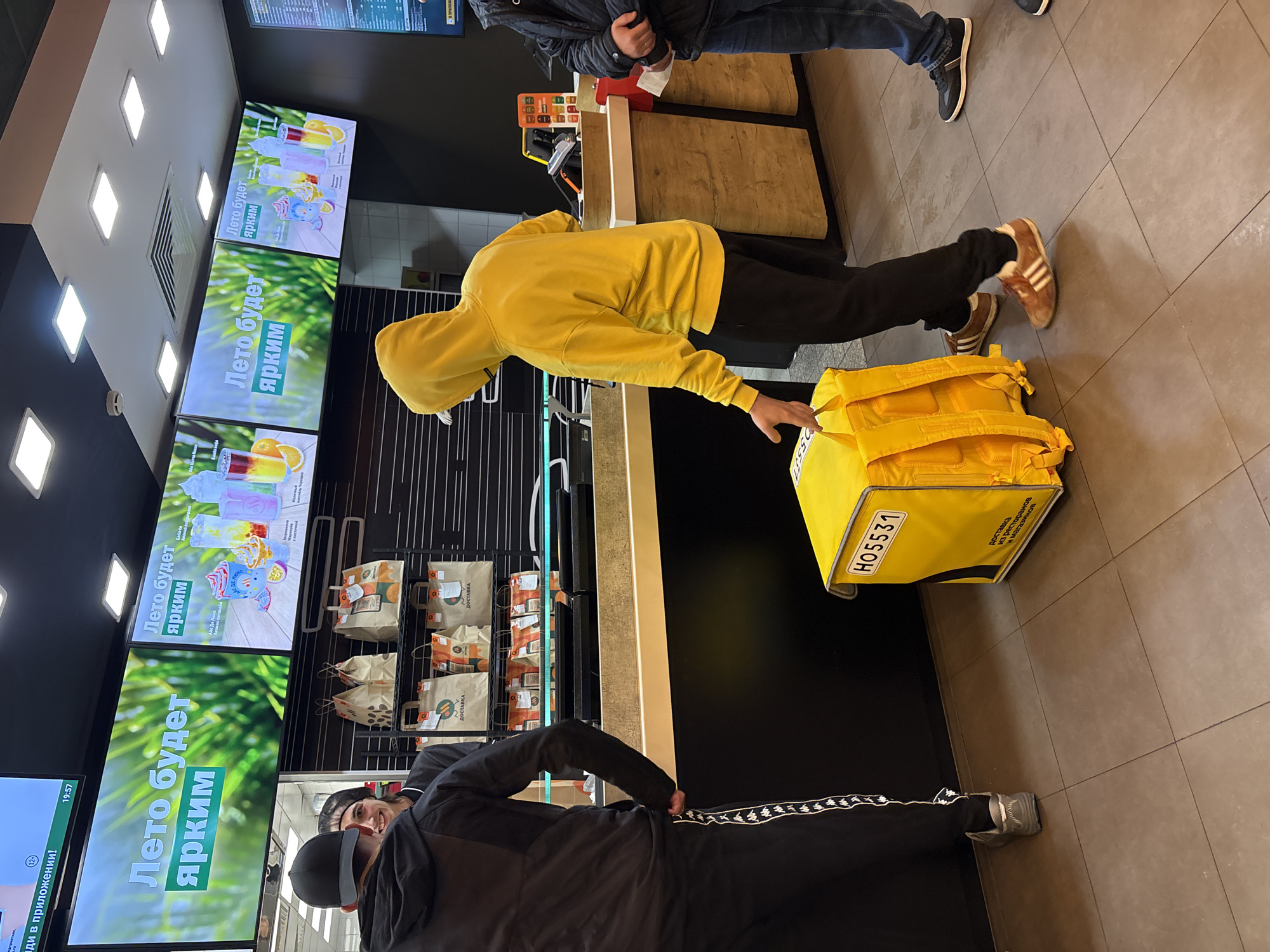 Here is a description of the image:
**Location:** A McDonald's restaurant in Izhevsk, Russia, as evidenced by the Russian text on the digital menu boards.
**People:**
* A person wearing a yellow hoodie and dark pants is leaning over a counter, with their hand near a yellow food delivery bag. They appear to be a delivery person picking up an order.
* Two other people are partially visible: one wearing a dark jacket and jeans and another wearing a dark jacket with white stripes on the pants.
**Objects:**
* A large yellow delivery bag with the number "H05531" visible.
* Digital menu boards with images of beverages and text in Russian ("Лето будет ярким" meaning "Summer will be bright").
* Paper delivery bags displayed behind the counter.
* Food preparation area behind the counter.
**Actions:**
* The person in the yellow hoodie is reaching toward the yellow bag. It is likely the delivery person picking up the delivery order.
* Other people are waiting in line.
**Environment:**
* A tiled floor.
* Modern interior with digital screens displaying menu items.
* Standard counter setup of a McDonald's restaurant.