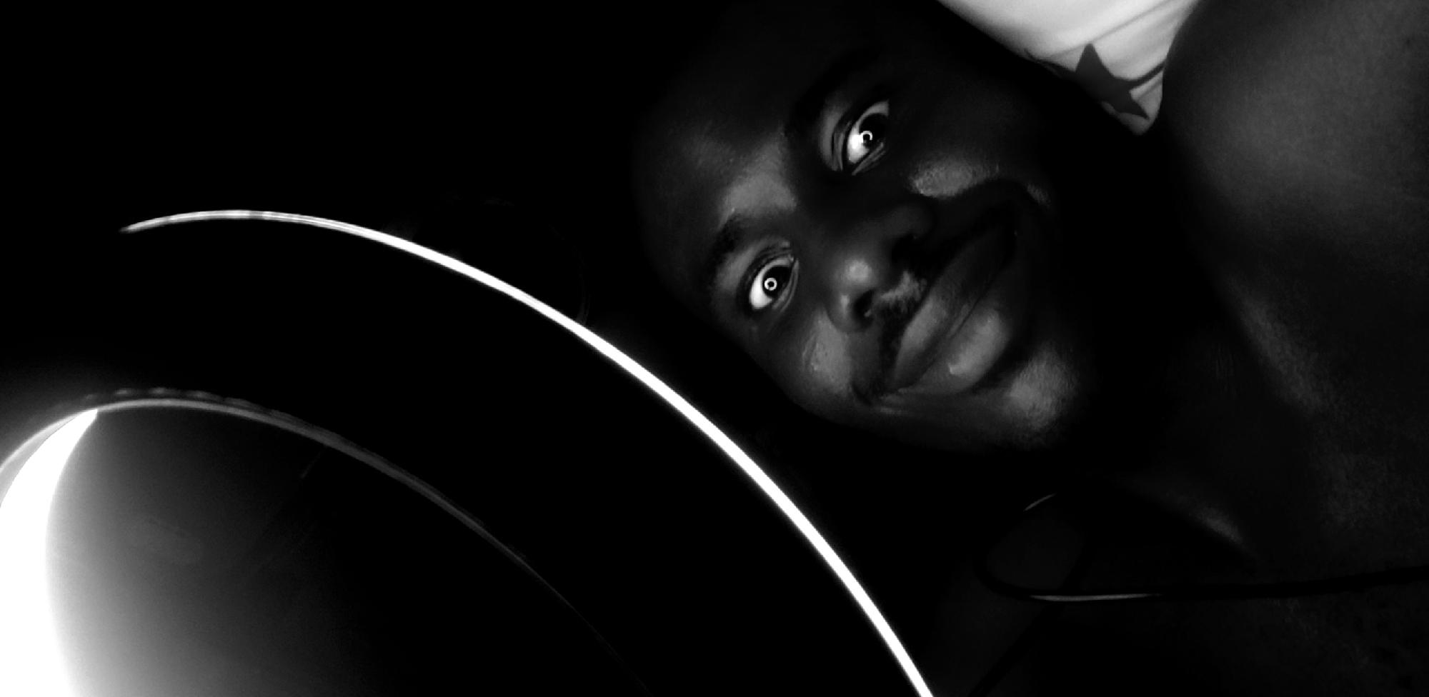 The image is a black and white, close-up portrait of a dark-skinned person with dark hair and a mustache. The person is lying down, with their face angled towards the camera. Their eyes are wide and appear to reflect light sources. The background is mostly dark, with a bright, circular light source visible in the lower-left corner, creating a bright arc shape. There is no contextual information in the image to definitively determine if it was taken in Narayi, Nigeria, or any other specific location.