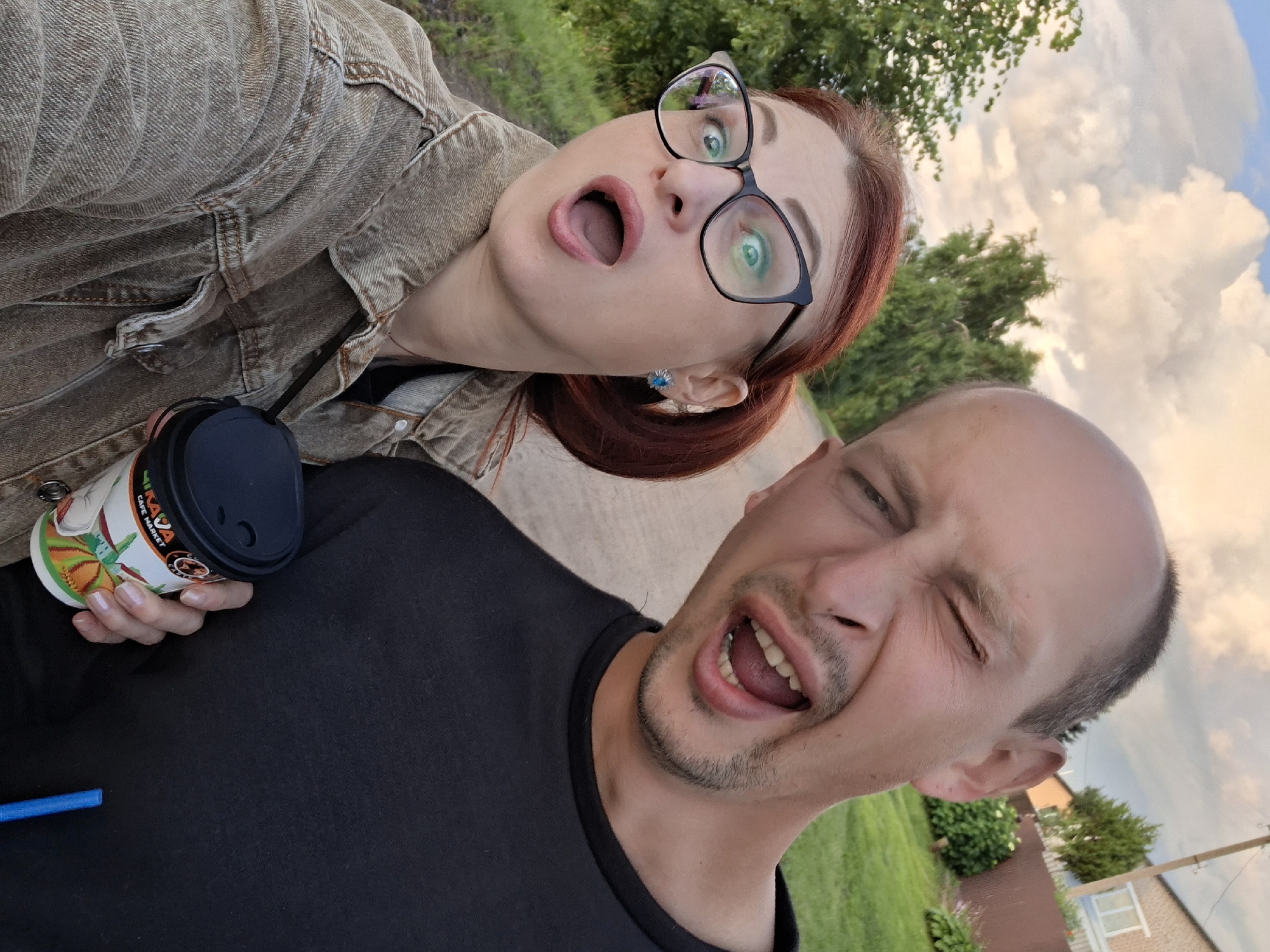 A selfie taken outdoors shows a man and a woman. The man, on the right, is balding with a mustache and goatee, and is winking with his mouth open. The woman, on the left, has red hair pulled into a ponytail, wears glasses, and has her mouth open in surprise. She is wearing a light brown denim jacket over a black shirt. The man is wearing a black t-shirt. The woman is holding a disposable coffee cup with a colorful design. A blue straw is visible next to the cup. The background suggests a rural or suburban setting with greenery and a building in the distance under a cloudy sky. This scene is located in the Zabalotski village council, Belarus.