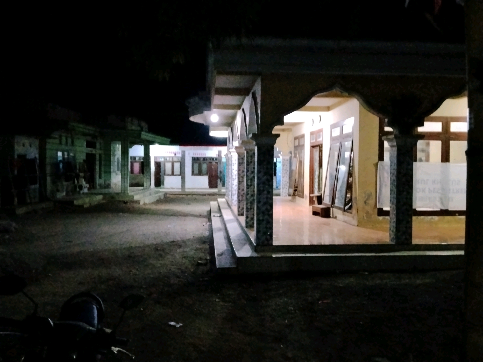 The image depicts a nighttime scene in front of a building that appears to be a village hall or community center in Panggungan, Indonesia. The foreground is dominated by the dark silhouette of a motorcycle, suggesting it is parked outdoors. The building itself is illuminated by artificial lights, casting a warm glow on the facade. The architecture features a covered walkway with decorative pillars and a raised entrance with steps. Through the windows and open doorways, interiors with white walls and wooden window frames are visible. The ground outside is unpaved and appears to be dirt or gravel. In the background, other structures with similar lighting and architectural elements can be seen, indicating a collection of buildings within the area. The overall atmosphere is quiet and still, typical of a rural Indonesian setting at night.