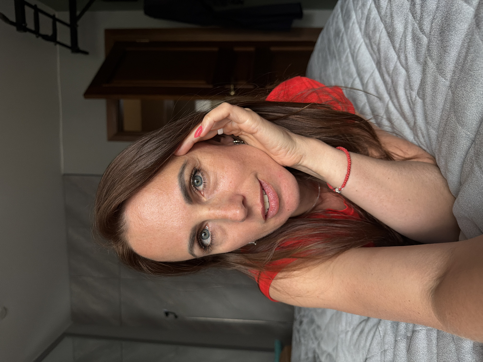 A woman with long brown hair and blue eyes is lying on a bed. She is wearing a red shirt. Her left arm is bent, and her hand is resting on her face, near her temple. A red bracelet is visible on her wrist. The background shows a bedroom with a closet, a door, and part of a wardrobe. The bed has a textured grey cover. There are no specific landmarks visible to indicate the city of Birobidzhan, Russia.