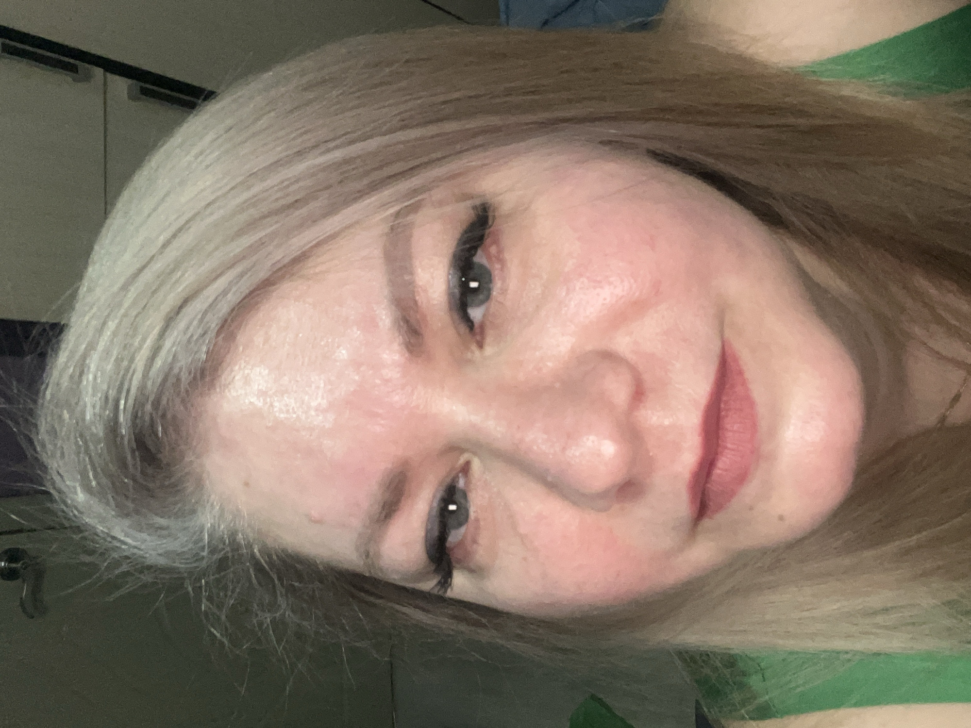A close-up, angled portrait shows a woman with light blonde hair. She has fair skin, dark eyeliner, and pink lipstick. Her expression is a slight smile. The background is indistinct, showing hints of interior objects. The location in Izhevsk, Russia is not discernible from the image.