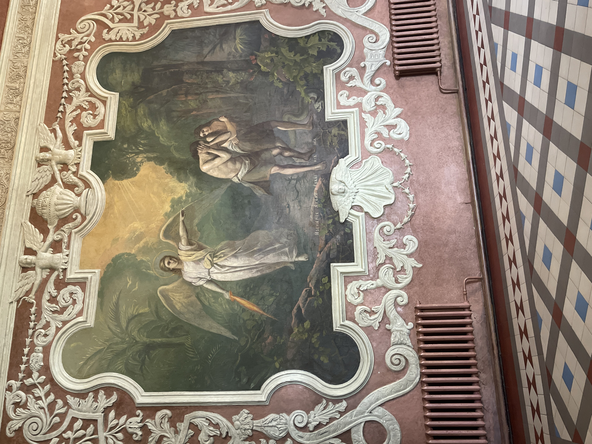 The image depicts a large, ornate mural painted on a pink textured wall. The mural is framed by elaborate white stucco ornamentation, featuring floral motifs and winged cherubic figures. The central painting depicts a biblical scene: an angel with a halo and a flaming sword stands to the left, pointing towards the right. To the right, a man and a woman, presumably Adam and Eve, are shown in a state of distress, with the woman embracing the man as he covers his face. A snake is visible at their feet. The background of the painting features lush greenery, trees, and a bright, sunlit sky. Below the mural, on the wall, are two large, industrial-style radiators. The floor in the foreground is tiled in a geometric pattern of red, gray, and blue squares. The scene is likely located in a public building, possibly a museum or a historical institution, given the grandeur of the mural and the architectural details. The text visible below Adam and Eve is in Cyrillic script. The overall impression is one of religious or historical significance, rendered with classical artistic style.