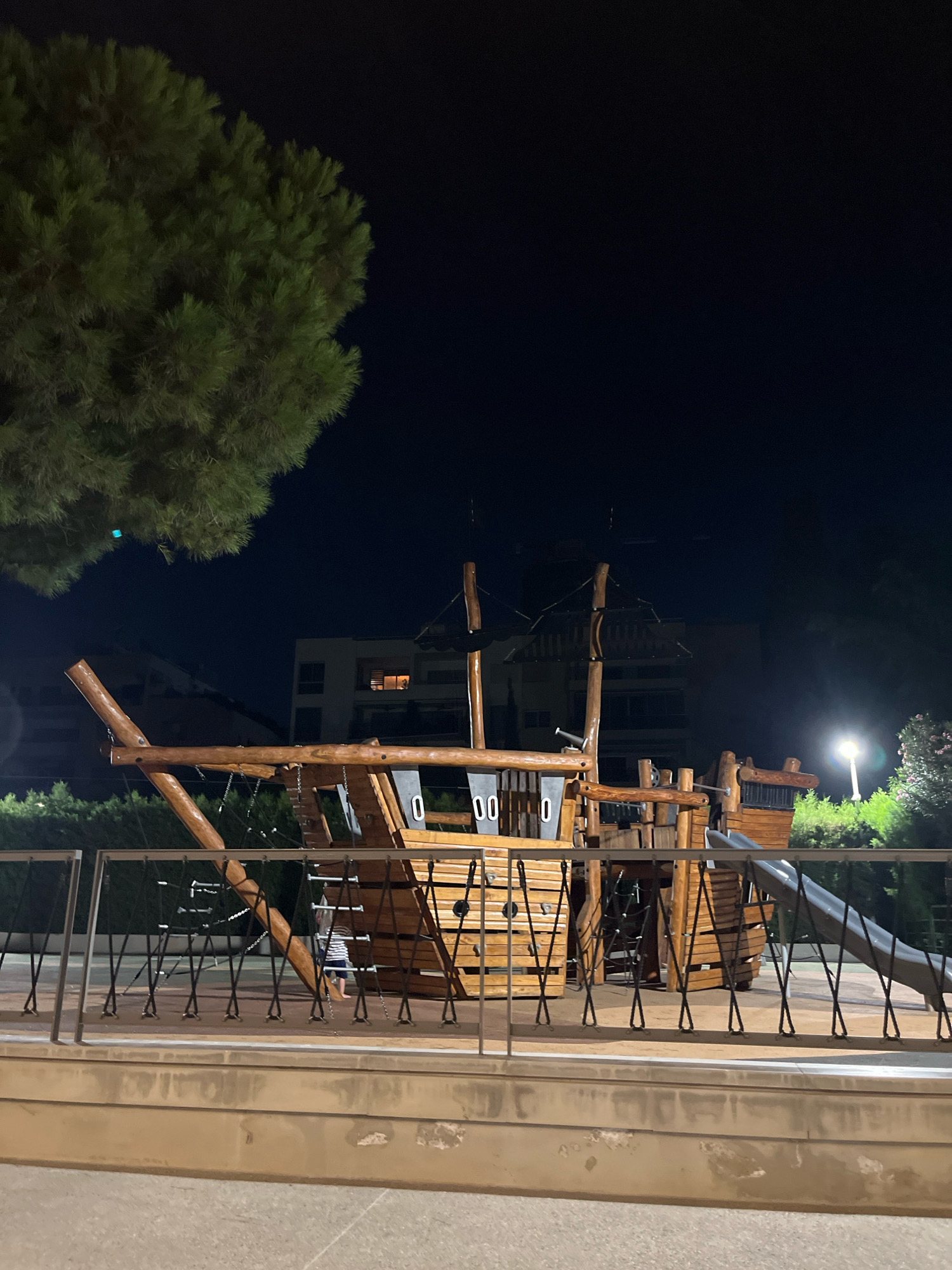 In Agios Tychona Community, Cyprus, a wooden ship-themed playground is visible at night. The playground features wooden structures resembling a ship, complete with masts, railings, and a slide. A child, wearing a striped shirt and shorts, is walking within the playground. A streetlamp illuminates the scene, casting a glow on the surrounding green bushes and trees. In the background, a multi-story building with illuminated windows is visible. The sky is dark, indicating nighttime.