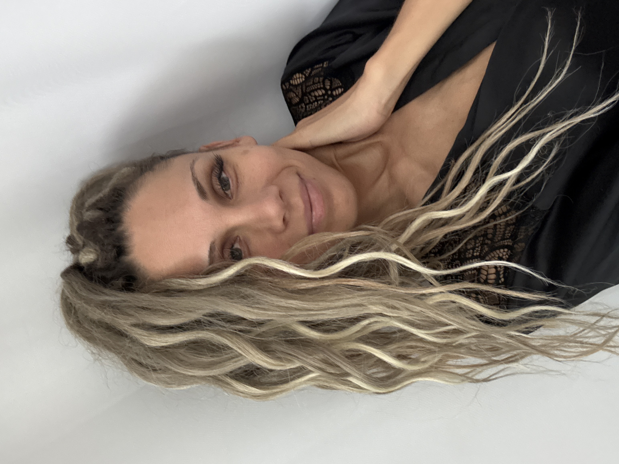 The image shows a woman with long, wavy blonde hair pulled up into a high ponytail. She is wearing a black robe with lace detailing on the shoulders and neckline. Her head is tilted and she is looking towards the camera with a slight smile. Her right hand is touching her neck. The background is a plain white wall. There is no indication of the city of Rusanovo, Russia.