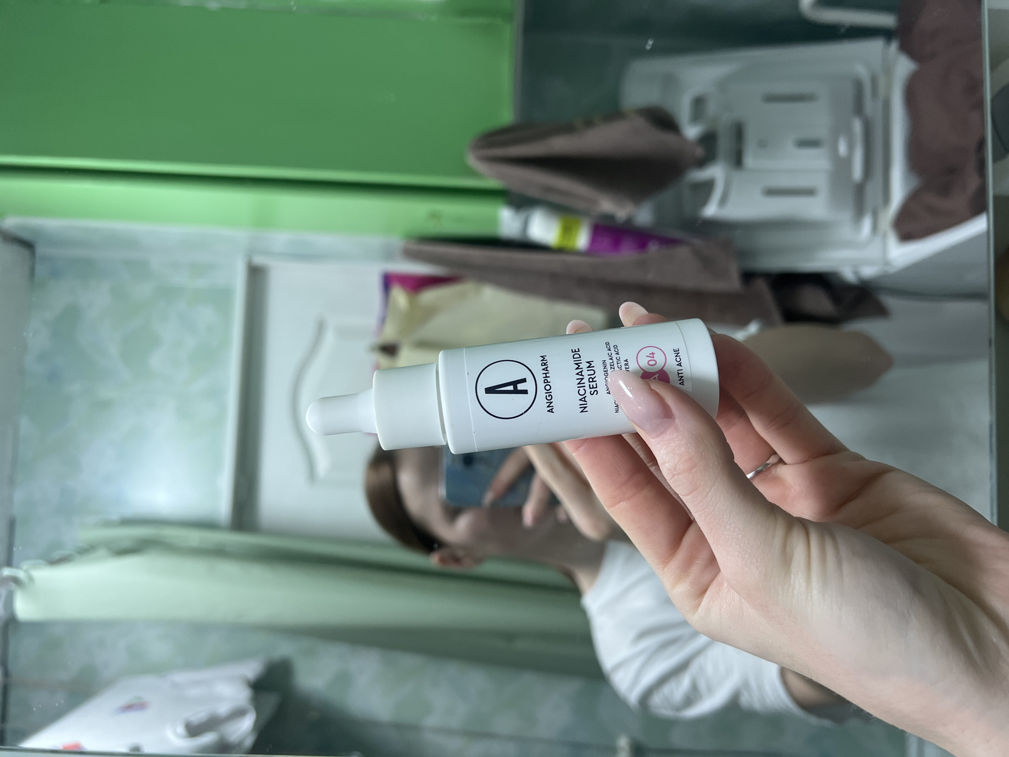 A person holds a white bottle of "Angiopharm Niacinamide Serum" in their hand, seemingly in a bathroom. The background shows a green surface, a rack with folded brown towels, and toiletries. The reflected image suggests the person is holding a phone, possibly taking a photo or video. While it's difficult to pinpoint the exact location as Chaikovsky, Russia, based solely on this image, the presence of typical bathroom fixtures and cosmetics indicates an indoor domestic setting.