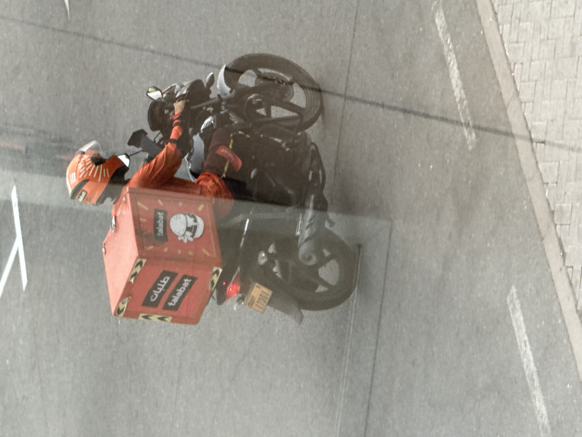 An overhead shot captures a delivery person on a motorcycle in Dubai, United Arab Emirates. The delivery person, wearing an orange helmet and an orange long-sleeved shirt, is riding a black motorcycle. Attached to the back of the motorcycle is a red delivery box with the word "talabat" written on it in both Arabic and English, along with a cartoon image of a burger. The motorcycle has a license plate that reads "17291". The scene takes place on an asphalt road marked with white lines, typical of a street in a city. The image appears to be taken through a window, as suggested by a faint reflection across the scene.