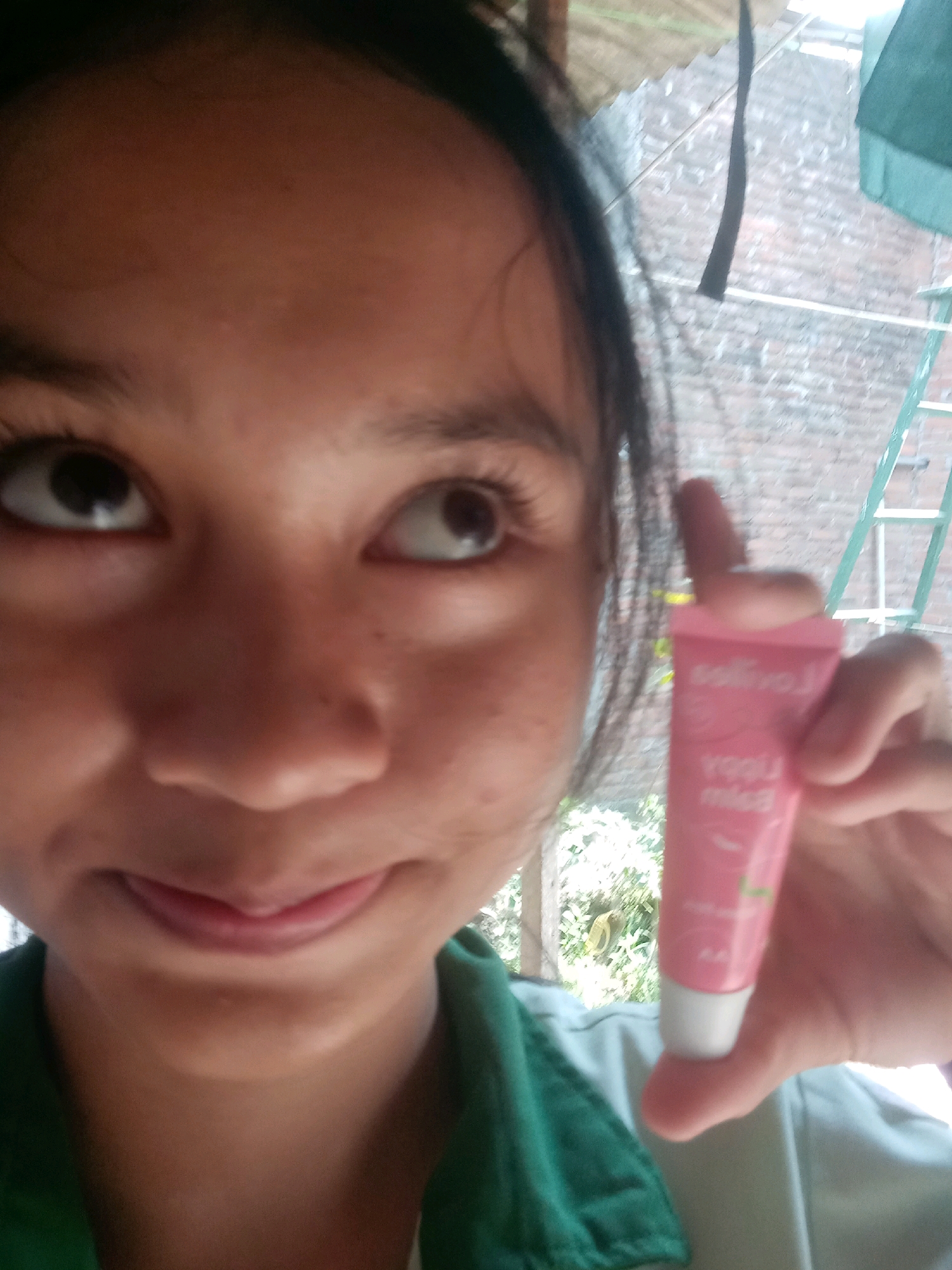 A young person, seen from the chest up, is holding a small, pink tube of lip balm to the camera. The person's face is slightly blurred, but they appear to be looking directly at the viewer with a faint smile. Their eyes are dark and wide, and their lips are partially closed, showing a hint of pink. The tube of lip balm has some text printed on it, which is partially legible as "misa."
The background suggests an outdoor setting, with greenery visible to the left and a brick wall and a ladder-like structure to the right. The lighting is somewhat diffused, hinting at either an overcast day or shaded conditions. The overall impression is casual and personal, like a selfie taken in a backyard or an outdoor common area. The presence of the lip balm suggests a moment of personal care or a demonstration of a product. The location context of Badas, Indonesia, combined with the visible elements, points to a residential or semi-rural environment.