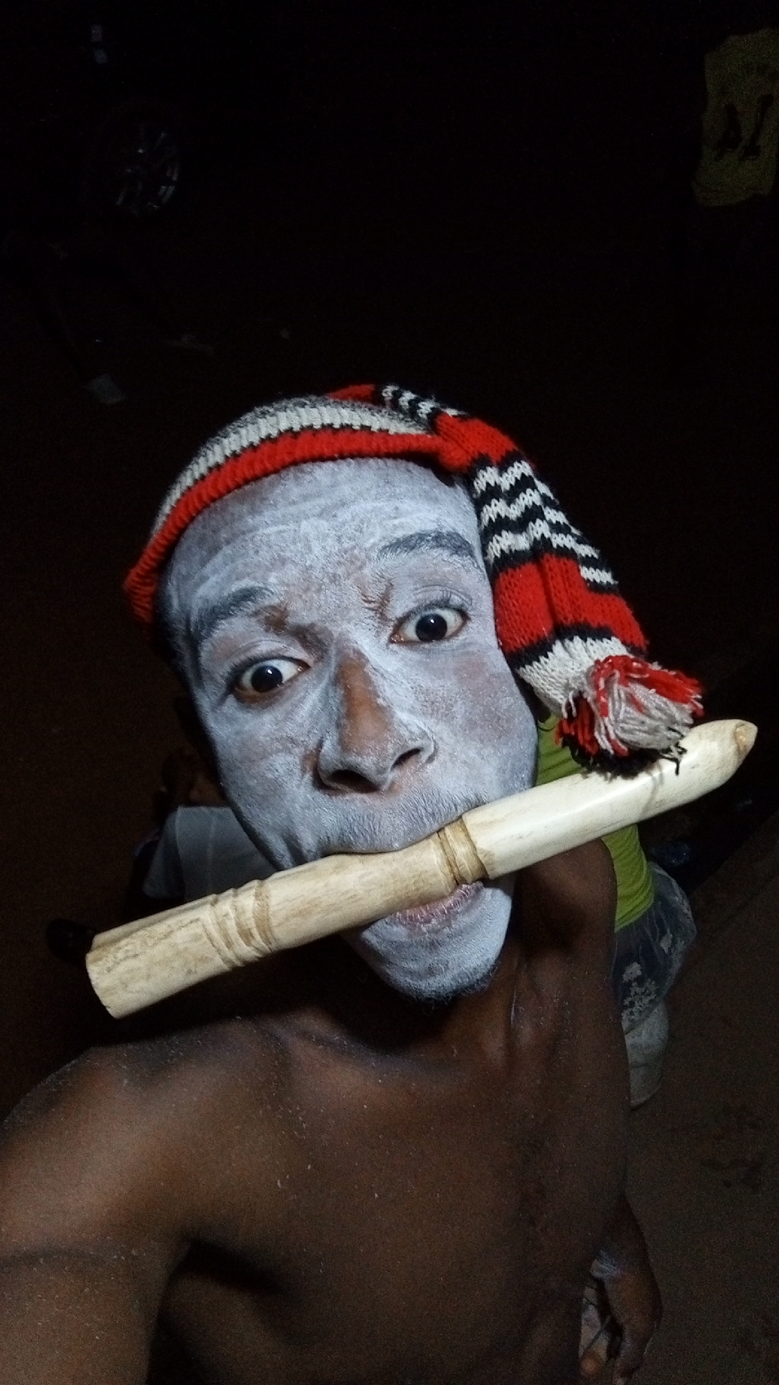 A dark-skinned person with white paint covering their face wears a striped knit cap and holds a light-colored, cylindrical object in their mouth. The environment appears dimly lit, with indistinct shapes in the background. The location is identified as the city of Onitsha, Nigeria.