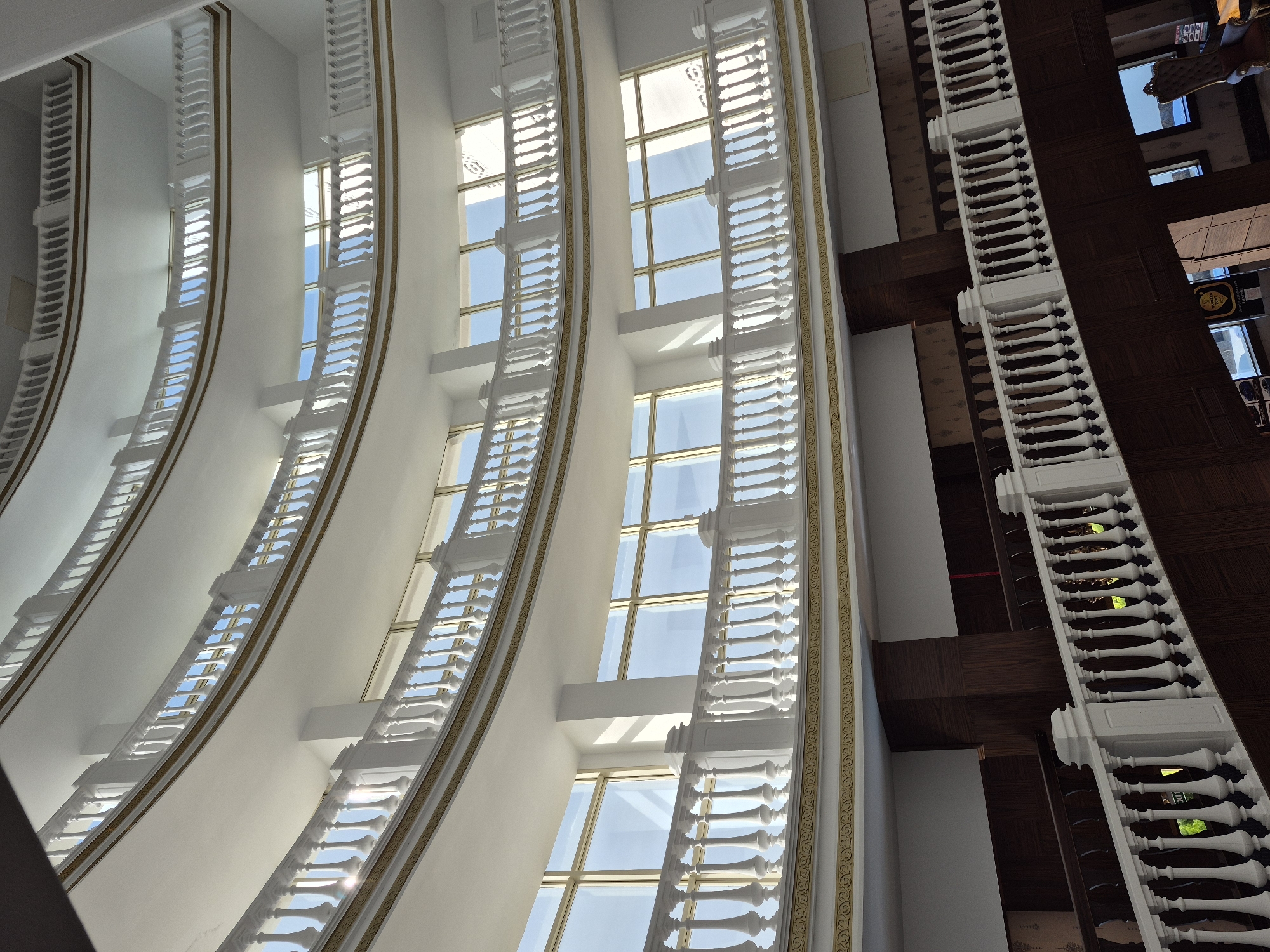 The image shows an interior view of a building in Alanya, Turkey. The architecture features a series of large, arched windows with ornate white balustrades and gold detailing. Sunlight streams through the windows, revealing a glimpse of the sky. To the right, a section of a dark wooden floor with a white balustrade suggests a balcony or upper level. The perspective is looking upwards, emphasizing the height and design of the building.