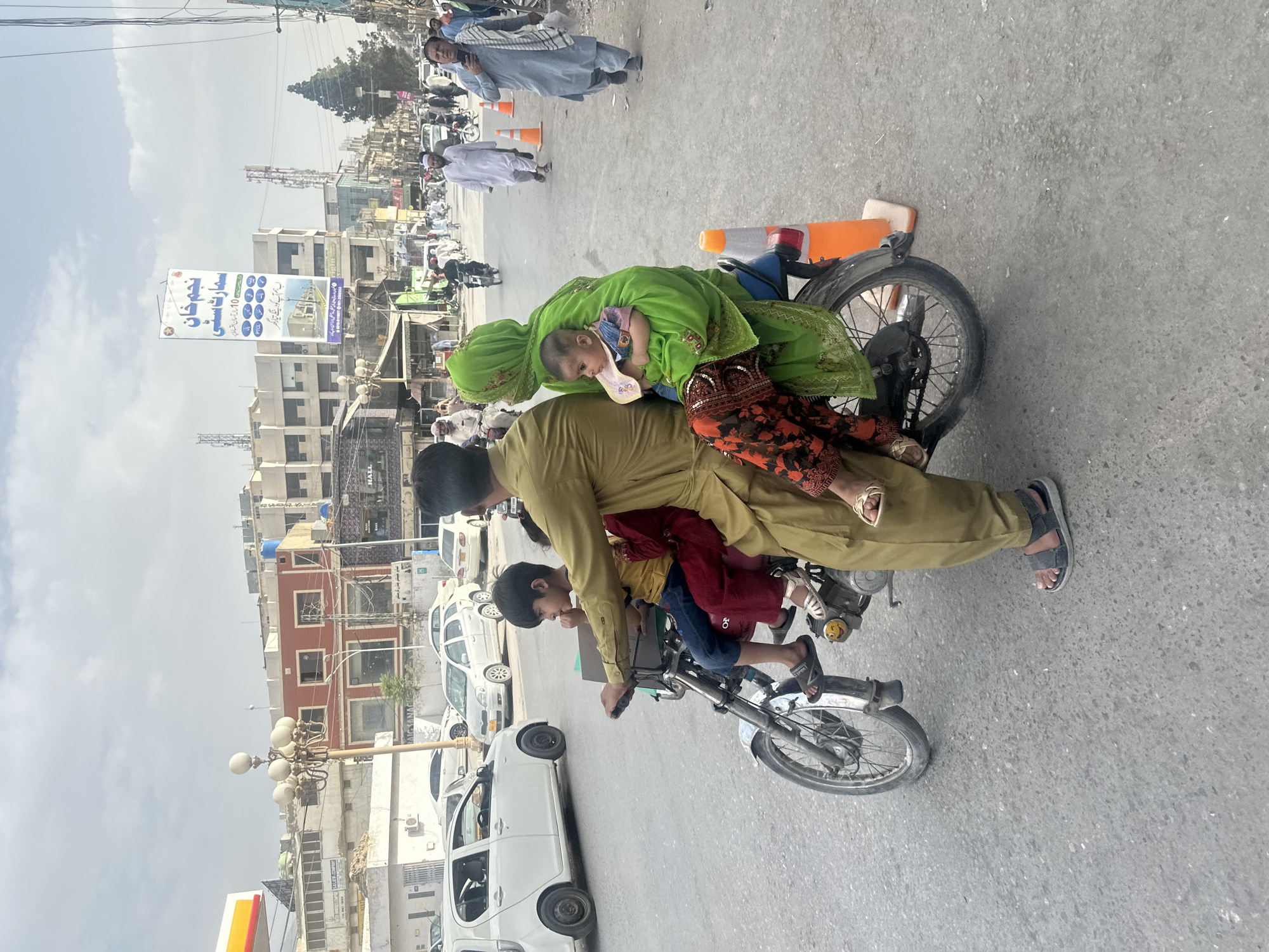 In Quetta City Tehsil, Pakistan, a family of four rides a motorcycle on a paved street. A man wearing a khaki outfit drives the motorcycle, with a child in a yellow shirt and blue jeans seated in front of him. Behind the driver, a woman in a green headscarf and dress holds a baby in her arms. The child in front is looking to the right and appears to be eating something. The woman is looking to the left, and the baby is looking straight ahead. The motorcycle is a red two-wheeled vehicle, with a silver fender and spokes on the wheels. Two orange traffic cones are visible behind the motorcycle. In the background, buildings line the street, some with signs in Urdu. Several cars and other motorcycles are also present on the street, and pedestrians are walking on the sidewalk. The sky is overcast with clouds.