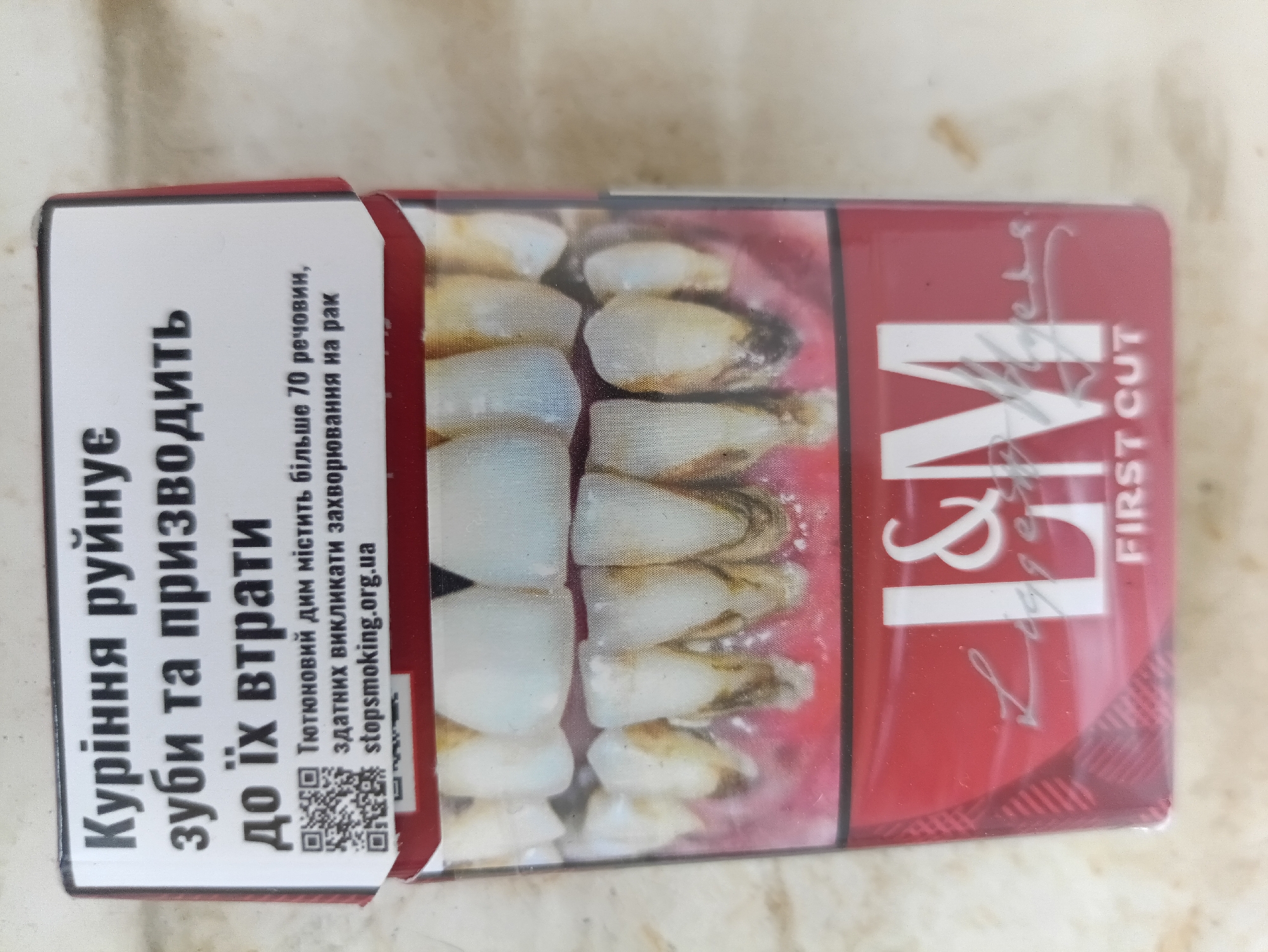 This media file is a close-up, top-down view of a pack of cigarettes. The pack is predominantly red and white, with a graphic image of unhealthy teeth on the front. The text on the pack is in Ukrainian and English. The Ukrainian text translates to "Smoking destroys teeth and leads to their loss. Tobacco smoke contains over 70 substances capable of causing cancer." The English text on the pack reads "L&M FIRST CUT". There is also a QR code and the website "stopsmoking.org.ua". The background appears to be a textured surface, possibly a table or countertop, with a light-colored, mottled pattern. The lighting suggests an indoor environment. The overall impression is a product packaging designed with a public health warning.
