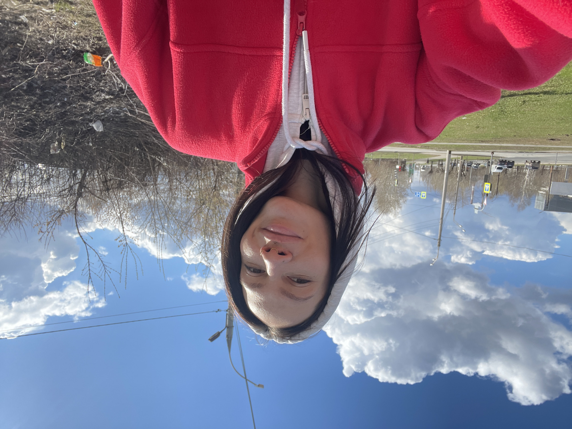 A young woman is captured in a selfie outdoors, looking directly at the camera with a gentle, pleasant smile. Her long, dark hair frames her face, and she has a small nose piercing. She is wearing a bright red fleece jacket over a light-colored hooded sweatshirt, the zipper of the jacket partially open.
The setting appears to be an urban or suburban area under a clear blue sky, adorned with prominent, fluffy white cumulus clouds. Bare tree branches, suggesting late autumn, winter, or early spring, are visible in the background, particularly to the left side of the frame. Beyond the trees, a road or street stretches into the distance, populated by cars, utility poles, and streetlights. Several yellow triangular traffic signs with black borders are discernible along the roadside, one appearing to signify a pedestrian crossing. Patches of green grass add to the outdoor scene.
A small, crumpled piece of litter, resembling a flag with distinct vertical stripes of orange, white, and green, is visible among the bare branches in the upper left of the background. The photograph conveys a bright, sunny day. No discernible text is visible within the image. The scene is located in Izhevsk, Russia.