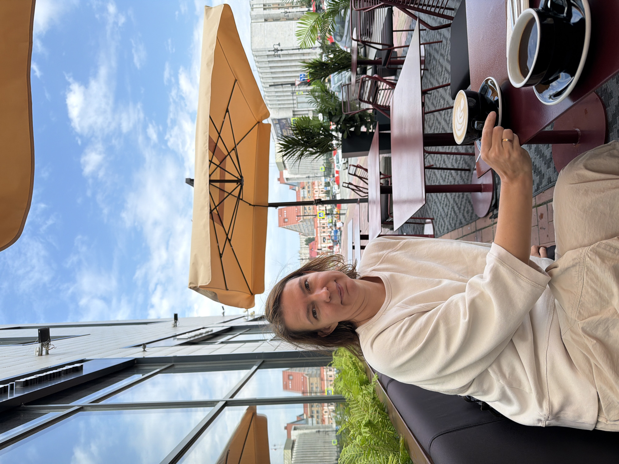 The image captures a woman sitting at an outdoor cafe in Perm, Russia. She is seated at a maroon table under a large, tan umbrella, with two black cups of coffee on the table. One cup contains a latte with latte art, and the other a darker coffee. The woman is wearing a light-colored sweatshirt and pants. In the background, there are other maroon tables and chairs, as well as potted plants. A modern building with large windows is to the left, reflecting the sky and surrounding cityscape. The street beyond the cafe shows traffic and buildings. The overall setting suggests a pleasant day at an urban cafe.