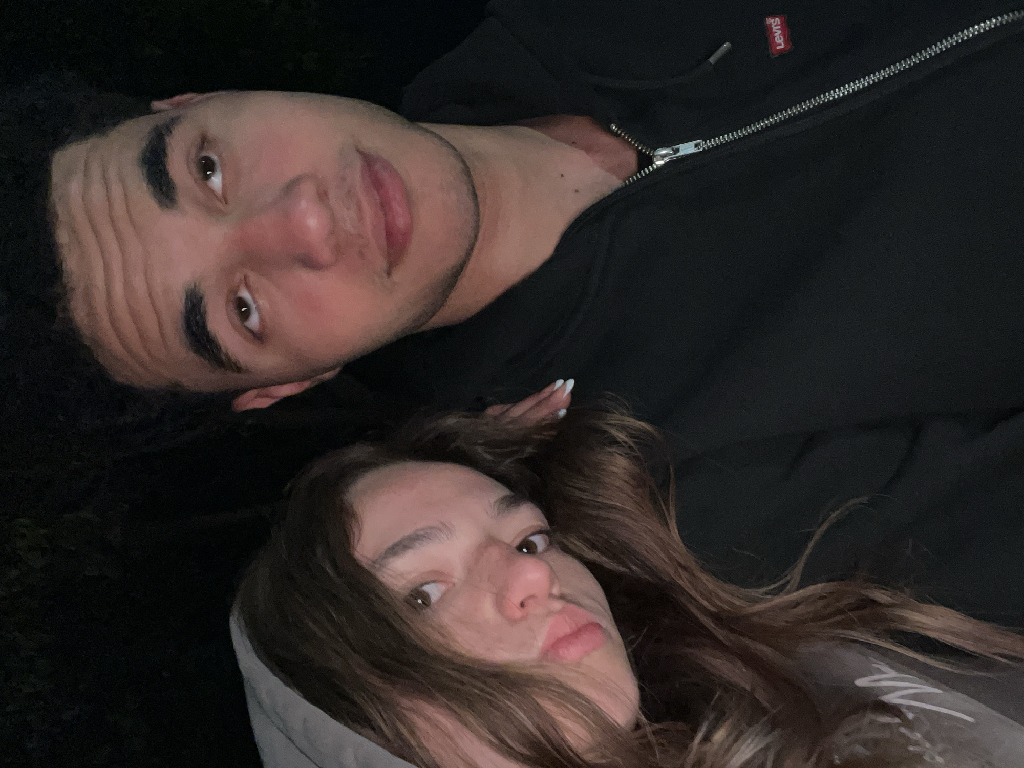 Two people, a man and a woman, are lying down. The man is wearing a black zippered jacket with a Levi's tag visible. The woman has long brown hair and is wearing a hooded sweatshirt with writing on it. They both appear to be looking at the camera. The location is not possible to determine.