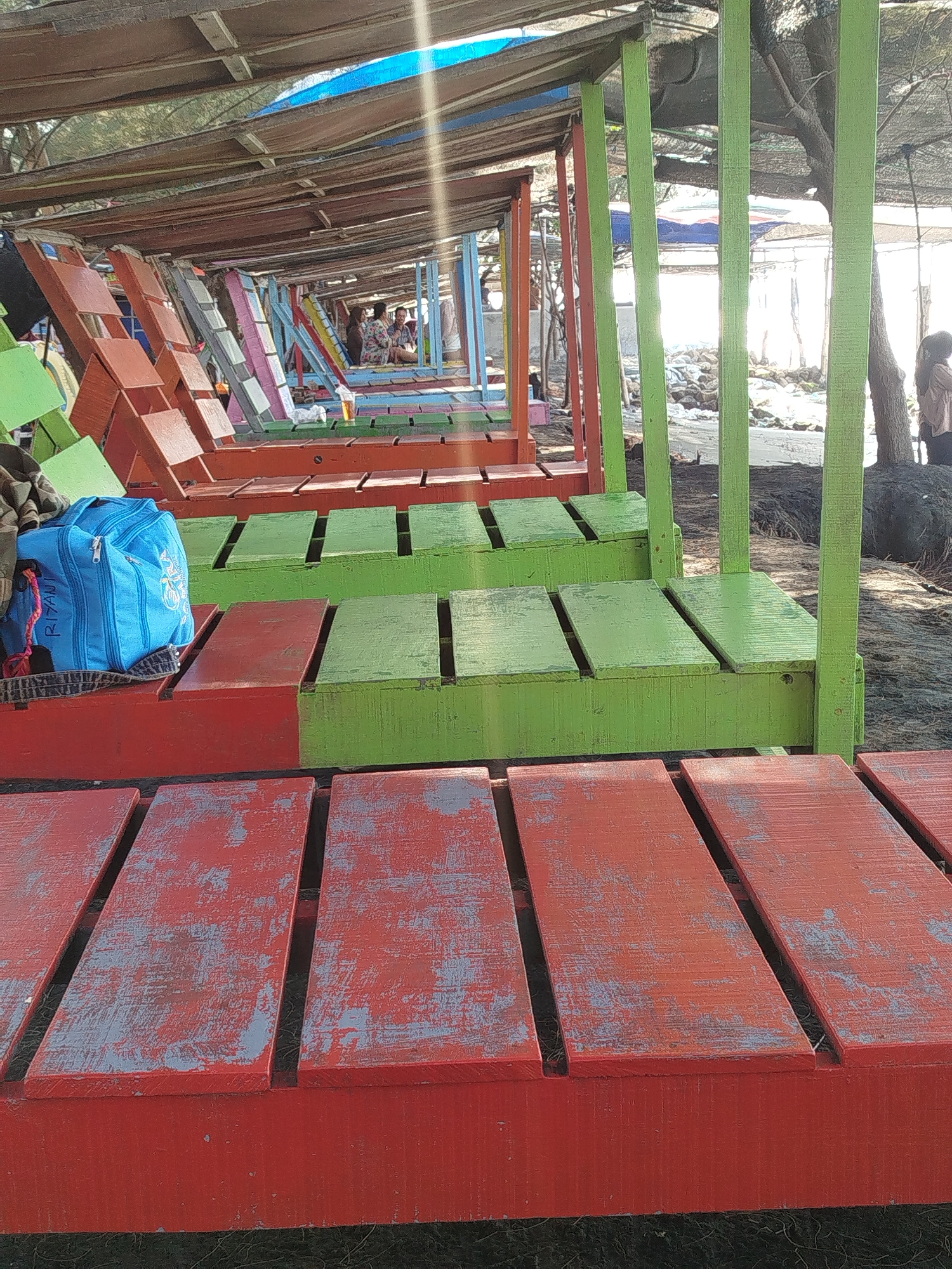In Mororejo, Indonesia, a beachside establishment features rows of colorful, weathered wooden seating. The foreground displays benches painted red with significant wear, showing glimpses of a gray undercoat. Behind these, green wooden benches are visible, also showing signs of age and use. Further back, a row of seating units transitions through various colors, including orange, pink, and blue. The seating appears to be arranged in a semi-covered area, with a wooden roof structure overhead supported by poles. In the background, a few individuals are seated at a table, and a blue backpack rests on one of the red benches in the middle ground. The environment suggests a casual, outdoor setting, likely a beach or waterfront area, with sand and natural vegetation present.