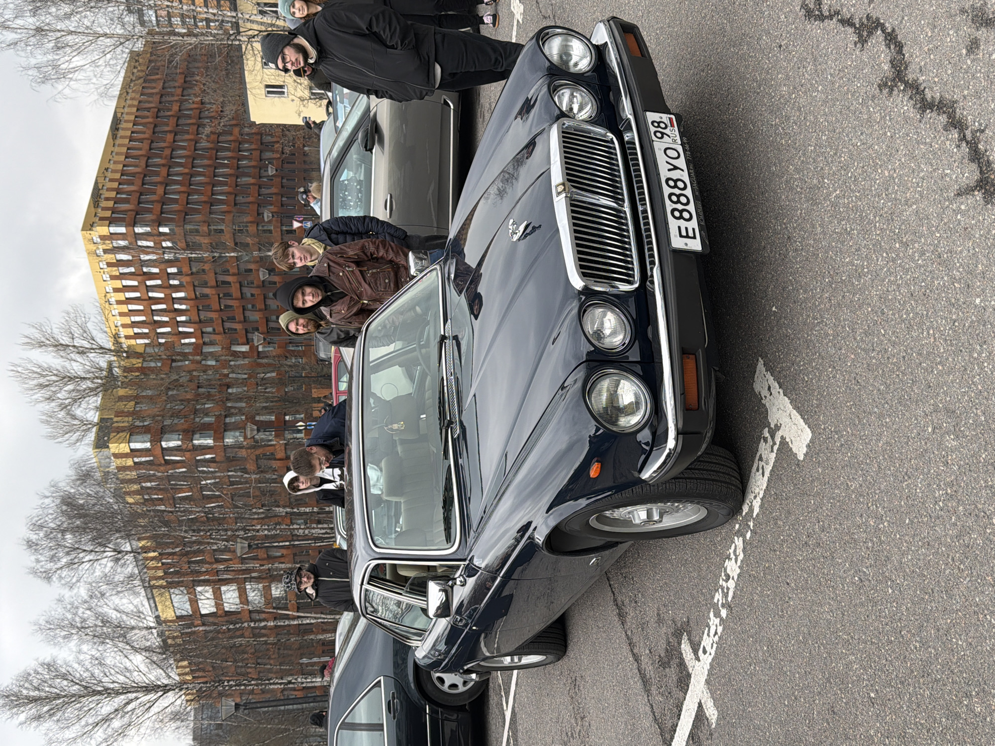 The image depicts a dark blue Jaguar XJ parked in Saint Petersburg, Russia. The car's front is angled towards the viewer, showcasing its distinctive grill, headlights, and the license plate "E888YO98" indicating its Russian registration. A group of people are standing nearby, some looking at the car, others at the camera. A modern building with a distinctive brick facade is visible in the background, along with bare trees suggesting a colder season. The car is parked on asphalt with marked parking lines. The overall setting is urban, with a mix of classic automotive design and contemporary architecture.