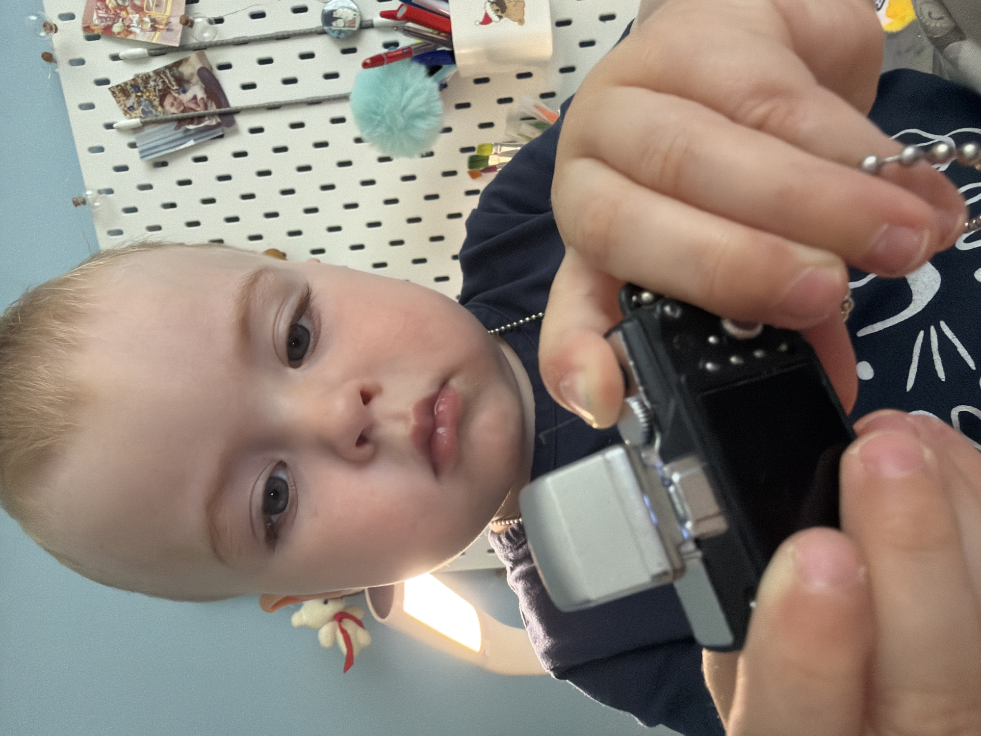 A fair-skinned young child with light blonde hair and dark eyes is the central focus of this close-up, sideways-oriented image. The child, dressed in a navy blue shirt with a white abstract pattern, holds a small black and silver miniature camera or toy attached to a silver beaded chain with both hands. Their expression is curious and direct as they gaze towards the viewer.
The background features a white pegboard filled with various personal items. Visible on the pegboard are small framed pictures, including one with a holiday theme, and another depicting what appears to be a child. A white container holds several pens, adorned with a small illustration of a bulldog wearing a Santa hat. A light blue fluffy pom-pom and miniature paintbrushes are also attached to the pegboard. To the bottom left of the child's head, a small white plush toy with a red ribbon hangs. A bright artificial light source is partially visible beneath the child's chin, casting some light upwards. The wall behind the pegboard is painted a light blue color.
The scene captures an intimate, candid moment indoors, likely in a child's room or play area, in Votkinsk, Russia. The child's activity revolves around exploring the small device, indicative of playful curiosity. No decipherable text is visible.