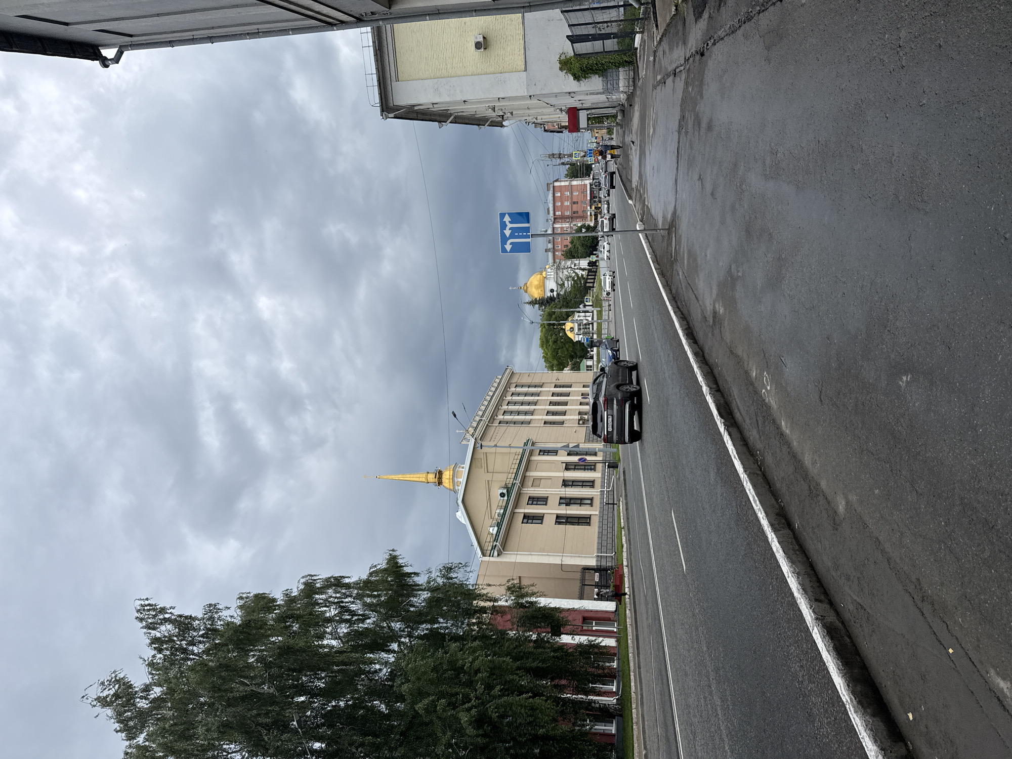 A street scene in Izhevsk, Russia, under a cloudy sky. On the left, a large building with a golden spire stands prominently, painted in light yellow and cream colors, with a row of windows. Adjacent to it, a darker building with red and white sections is partially visible. A large tree with dark green foliage is situated to the left of these buildings. The street in front is asphalt with white lane markings and a white-painted curb. A dark-colored SUV is driving on the road. On the right side of the street, a sidewalk made of asphalt is wet, indicating recent rain or moisture. A multi-story building, painted in yellow with white trim, runs along the right side of the sidewalk. In the distance, several other buildings, including a structure with golden domes, can be seen. Traffic signs, including one with directional arrows, are present along the street. A few pedestrians are visible, including one on the sidewalk and another walking on the street. The overall environment suggests an urban setting with a mix of architectural styles.