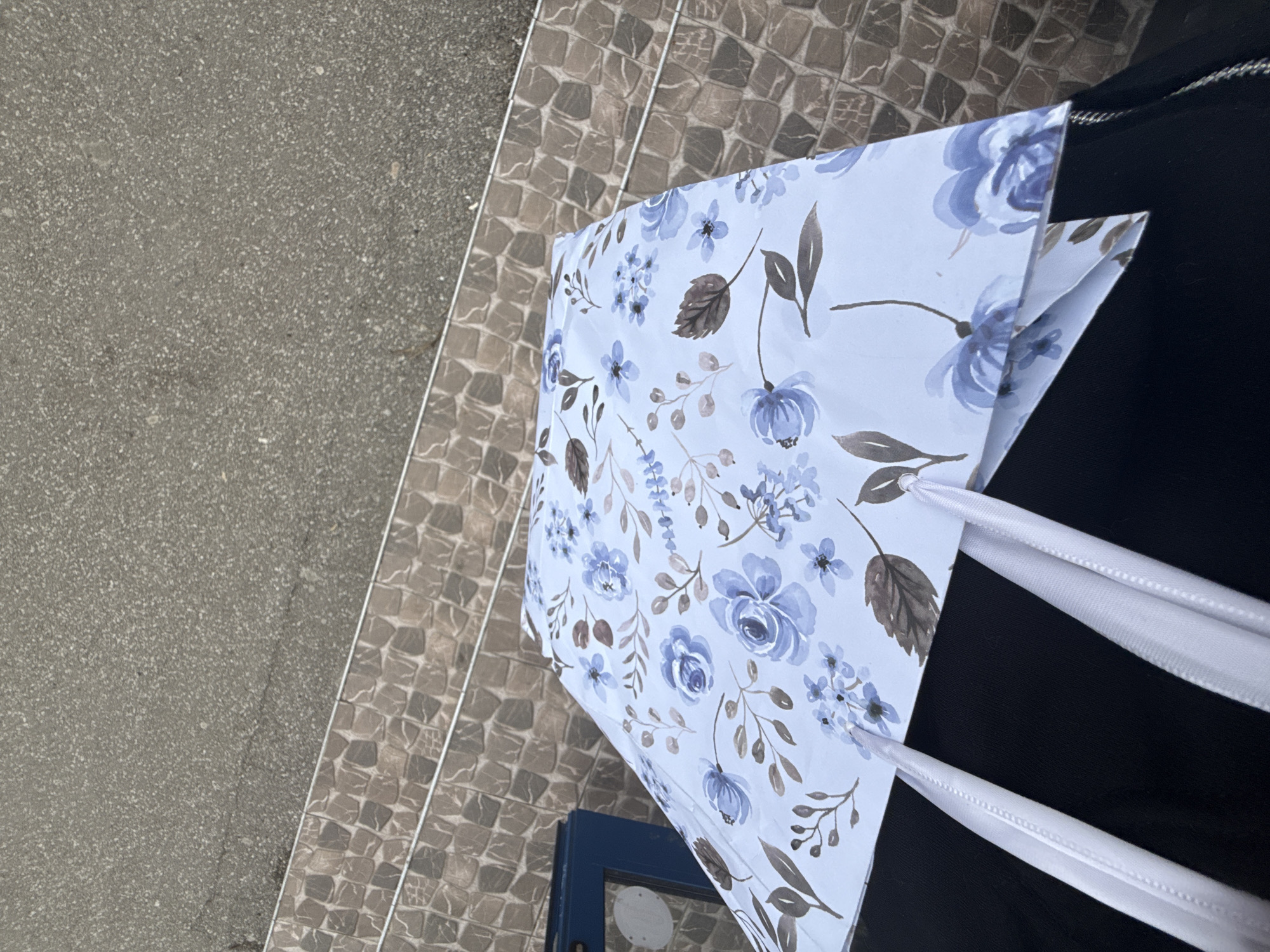 The image shows a gift bag with a floral pattern in shades of blue and brown. The bag has white ribbon handles. It is placed on a black surface, possibly fabric. The background consists of grey paving stones and stairs. The location is unspecified, but the city is identified as Sofia, Bulgaria.
