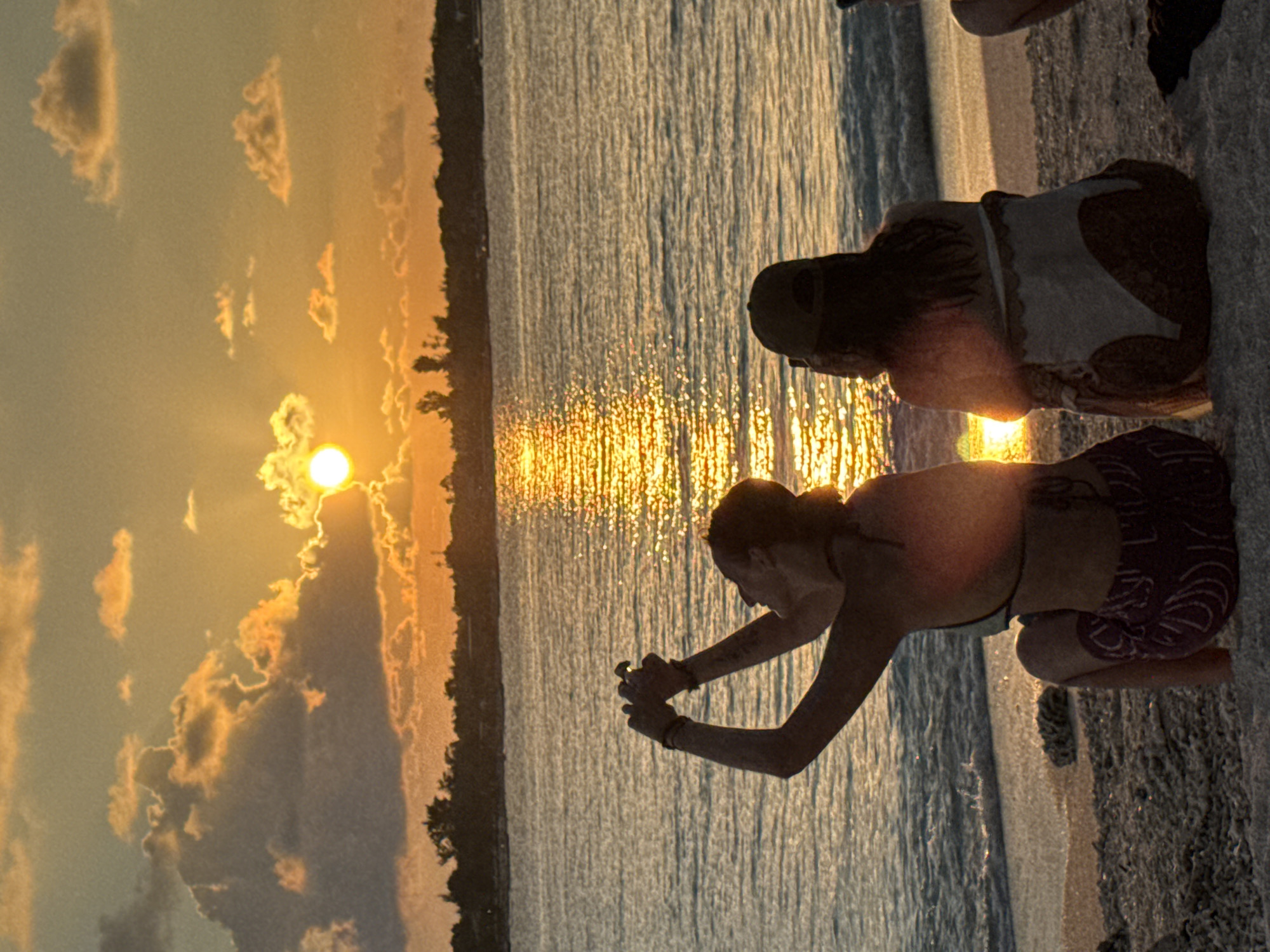 The vertically oriented image captures a serene golden hour scene at a shoreline, depicting either sunrise or sunset. A bright, low sun on the horizon casts a dazzling golden reflection across a body of water.
Two individuals are visibly kneeling or crouching by the water's edge. In the foreground, one person is seen in silhouette and partial back profile, wearing a bikini top and patterned shorts. Their arms are extended forward and slightly downwards, with hands clasped, suggesting an interaction with the water or an object at its surface. Further back, a second person is also kneeling, partially obscured, facing the water. The legs of a third individual are visible at the far right edge of the frame, hinting at more people present.
The expansive body of water, possibly an ocean or large lake, shimmers intensely with the reflected sunlight, creating a vibrant pathway of light towards the horizon. A dark, distant landmass or tree line is silhouetted against the bright sky. The sky itself is a blend of soft blues, oranges, and yellows, adorned with scattered, illuminated clouds.
The overall atmosphere conveys a sense of peaceful observation, contemplation, or appreciation of nature. The time of day is unequivocally sunset or sunrise, indicated by the sun's position and the warm, intense lighting. No visible text is present in the image. While the exact location is unknown from the image itself, the context suggests it is in Indonesia.