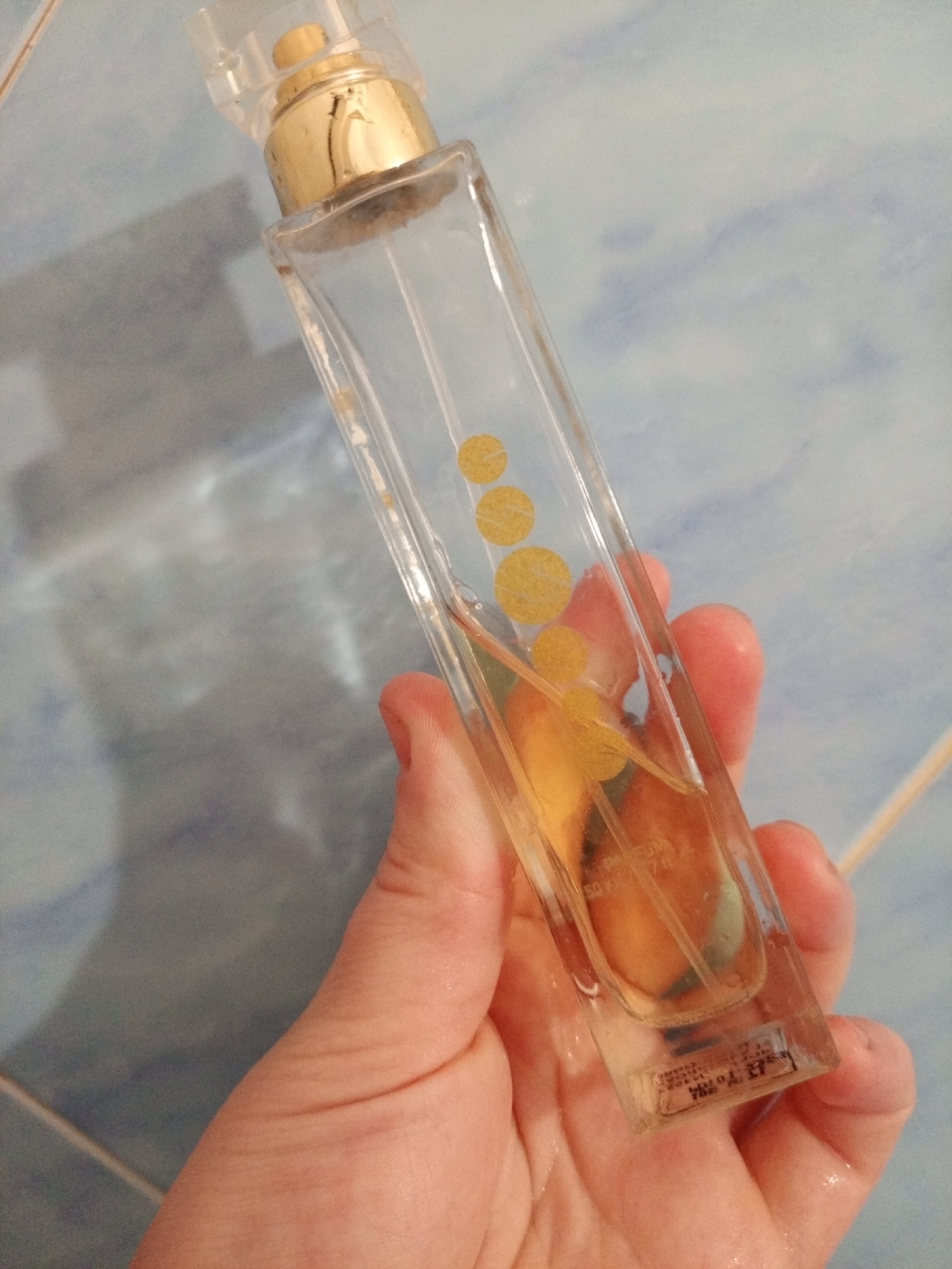 A hand holds a rectangular, clear glass perfume bottle with a gold-colored nozzle and a transparent cap. The bottle is partially filled with a light brown liquid. There are four gold-colored circles arranged vertically on the front of the bottle. The background is a light blue and white tiled surface. The location is Unknown, Unknown.