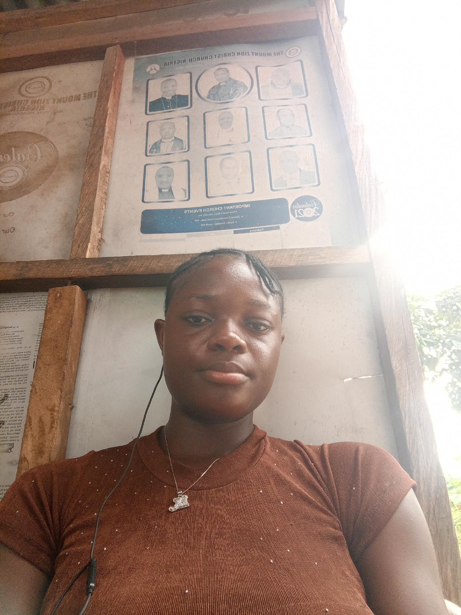 The photograph depicts a woman with dark skin wearing a brown shirt and a necklace with a pendant. She is seated in what appears to be an outdoor or semi-outdoor setting. Behind her is a wooden structure with two posters. One poster features a list of individuals, presumably church leaders, along with the text "THE MOUNT ZION CHURCH NIGERIA" in reverse. The other poster contains text and other information. Based on the limited information, a precise determination of the location within Mbribit, Nigeria, cannot be made.