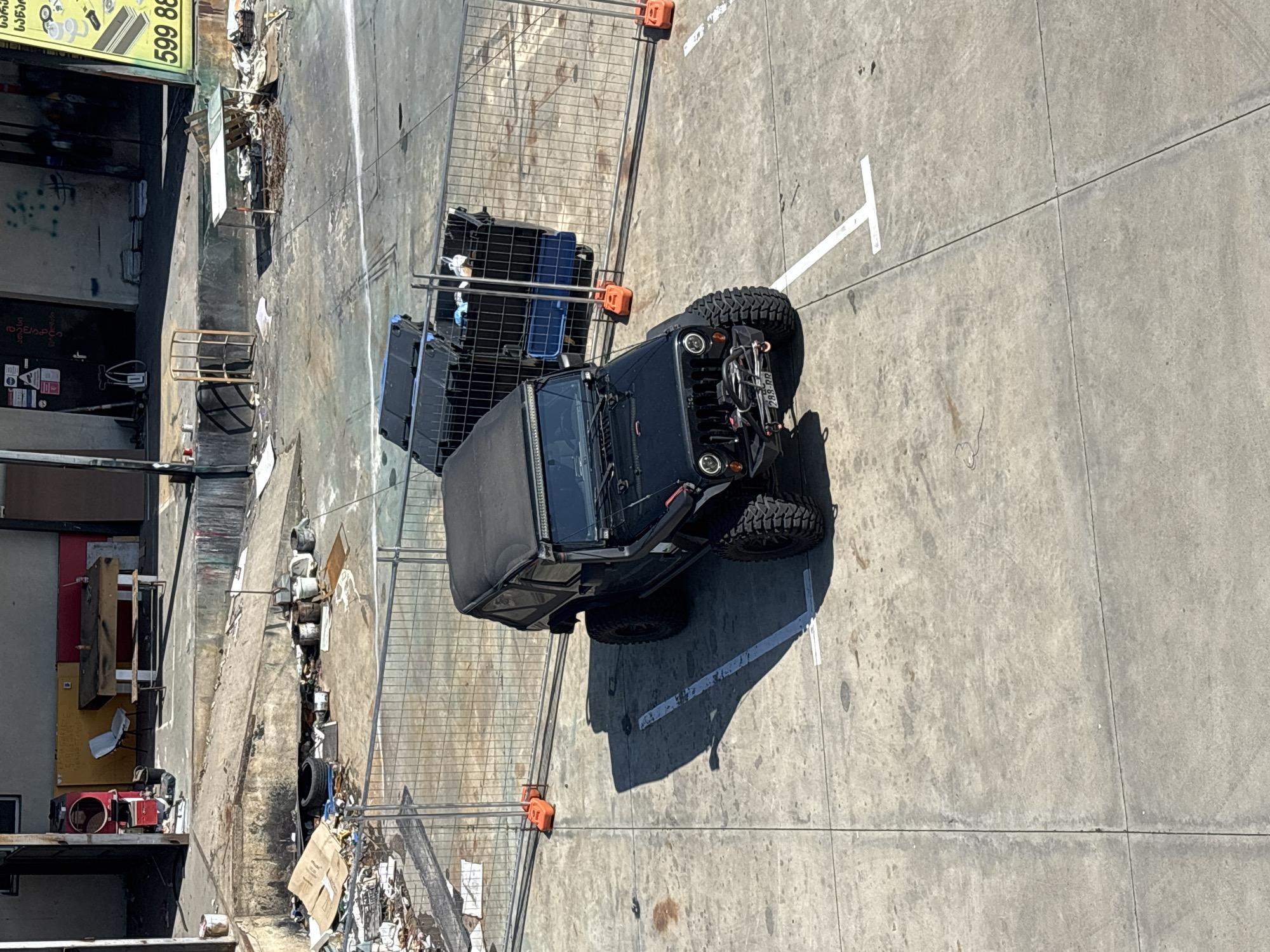 A black Jeep Wrangler is parked in a lot in Tbilisi, Georgia. The Jeep is facing right and is parked in a parking space marked by white lines on the concrete ground. The Jeep has a soft top and a light bar above the windshield. The surrounding area appears to be under construction or in a state of disrepair, with piles of debris, construction fencing, and scattered building materials visible. In the background, a building with a light-colored facade is visible, with some graffiti on its walls. The weather appears to be sunny, casting shadows on the ground.